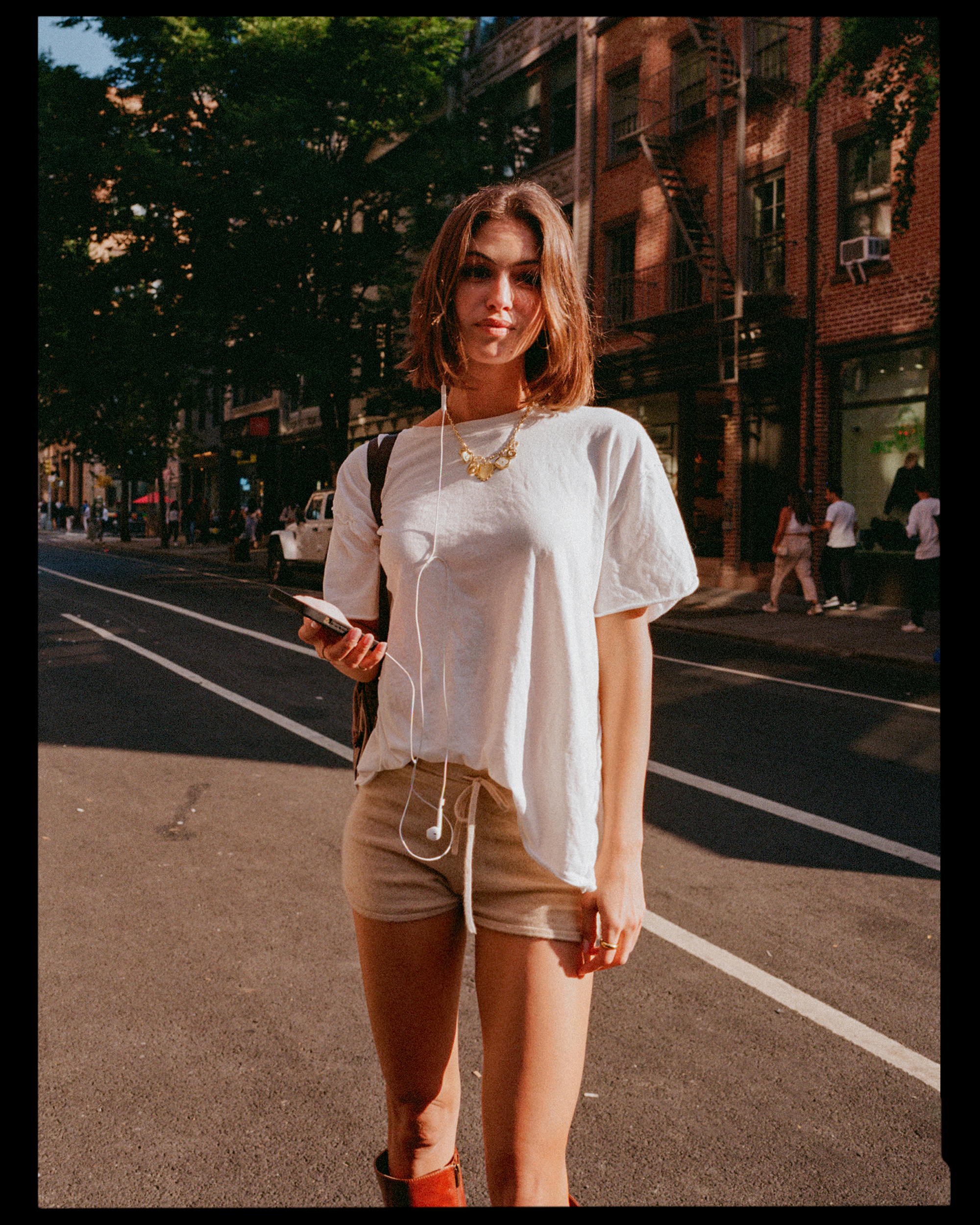 A young woman with short brown hair standing on a city street, holding a phone and wearing earphones, a white T-shirt, beige shorts, and brown boots, with a brick building, trees, and pedestrians in the background.