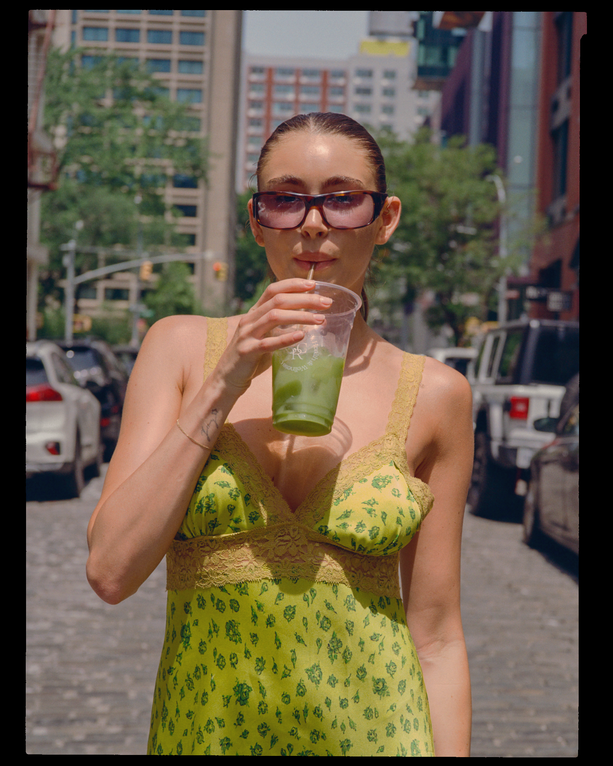 Woman wearing sunglasses and a yellow dress with a green floral pattern, sipping a green drink through a straw in an urban outdoor setting with buildings and parked cars in the background.