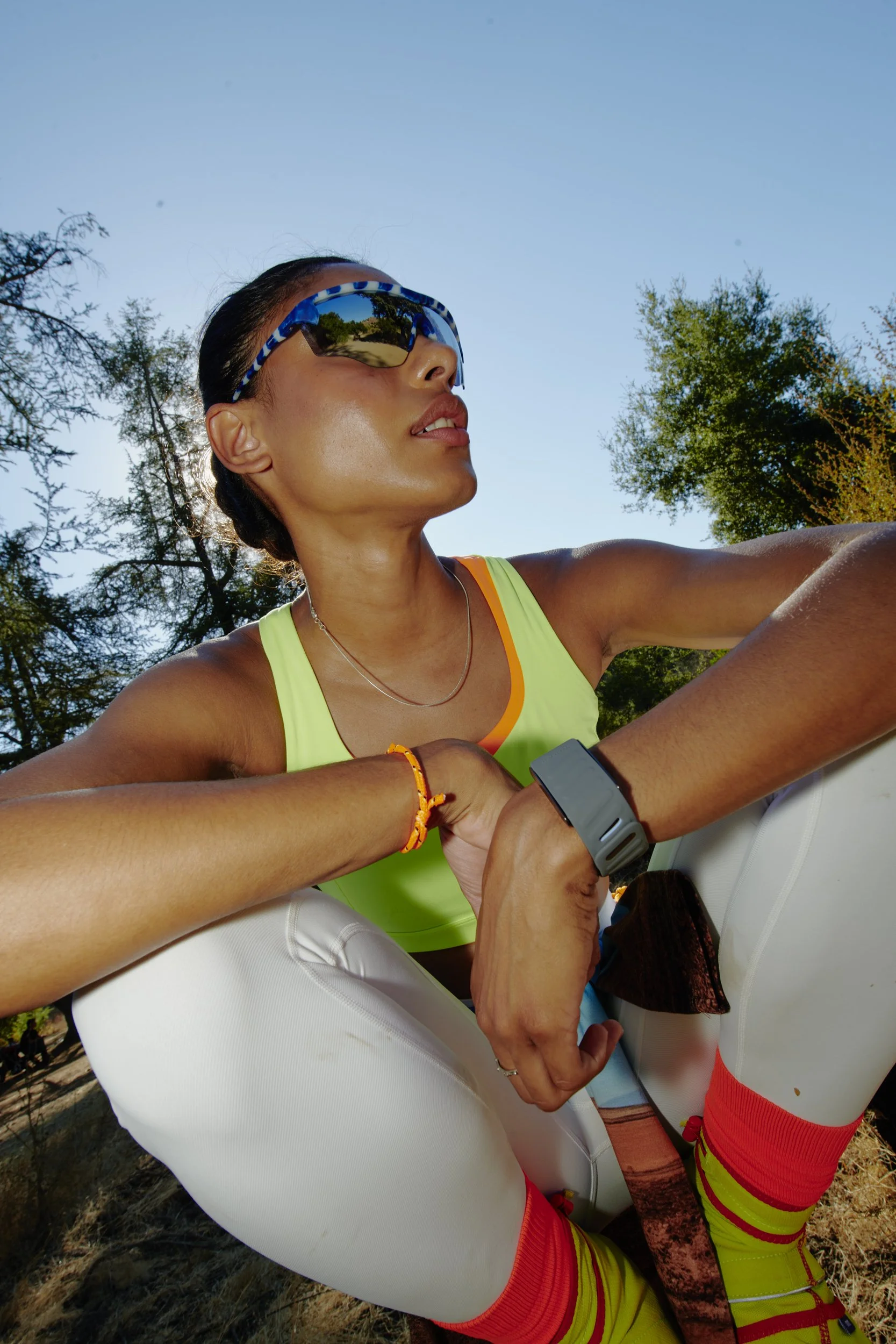 A woman with athletic build, wearing sunglasses, a yellow sports top, and blond hair tied back, is outdoors with trees in the background, posing for a selfie with her eyes closed.