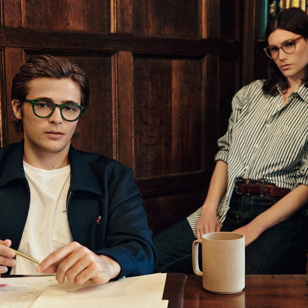 Two people sitting at a table in a wood-paneled room. The person on the left is a young man wearing glasses, a dark jacket, and a white shirt, holding a pencil and looking toward the camera. The person on the right is a young woman with glasses, wearing a striped shirt, sitting with her arm resting on the table, with a beige mug in front of her.