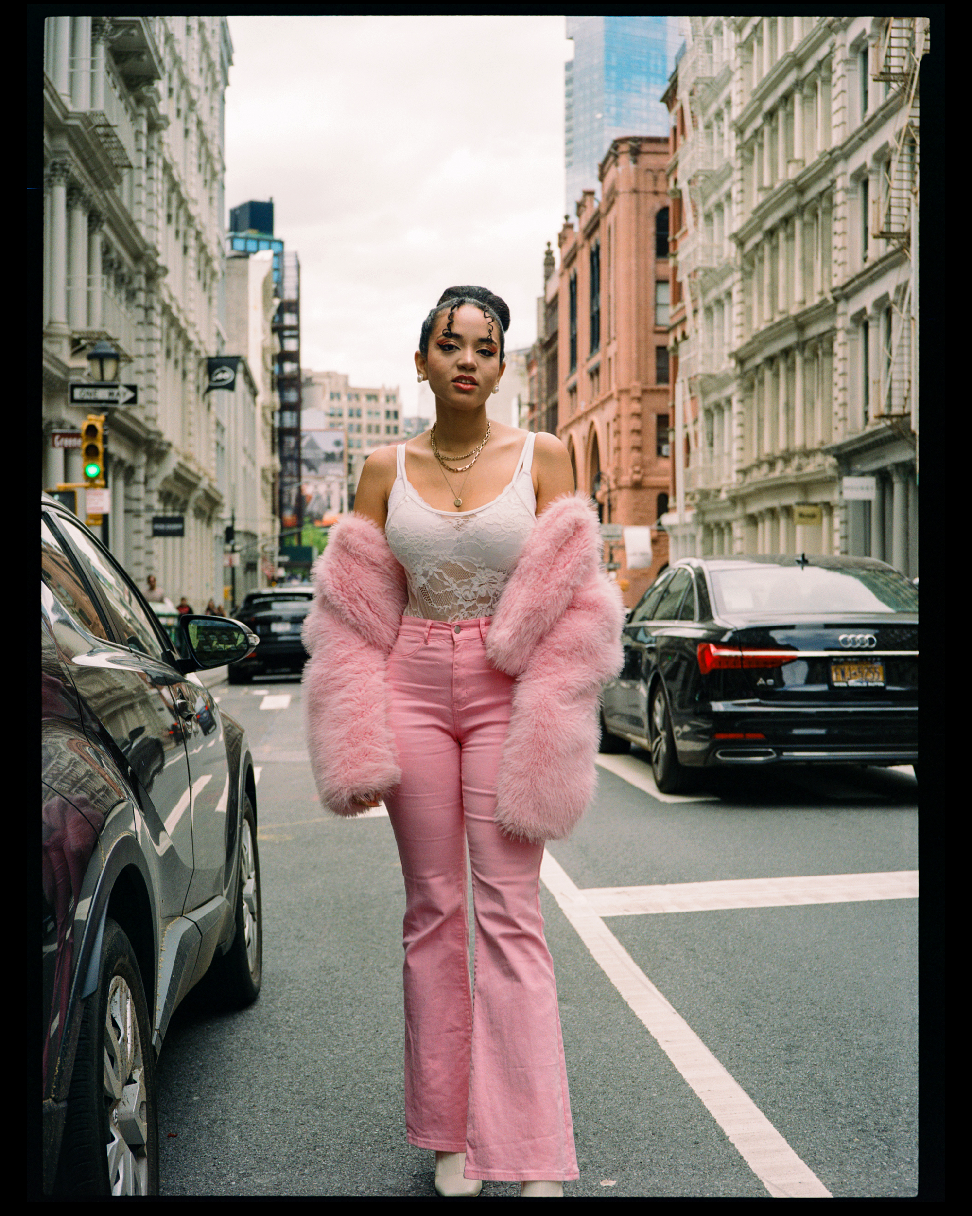 Young woman standing in the middle of a city street, surrounded by parked cars and tall buildings, wearing pink pants, a white lace top, a pink fuzzy coat, and jewelry, with a confident expression.