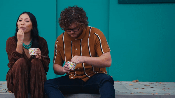 A woman and a man sitting against a teal wall, with the woman holding a colorful cup and the man scooping ice cream from a small container.