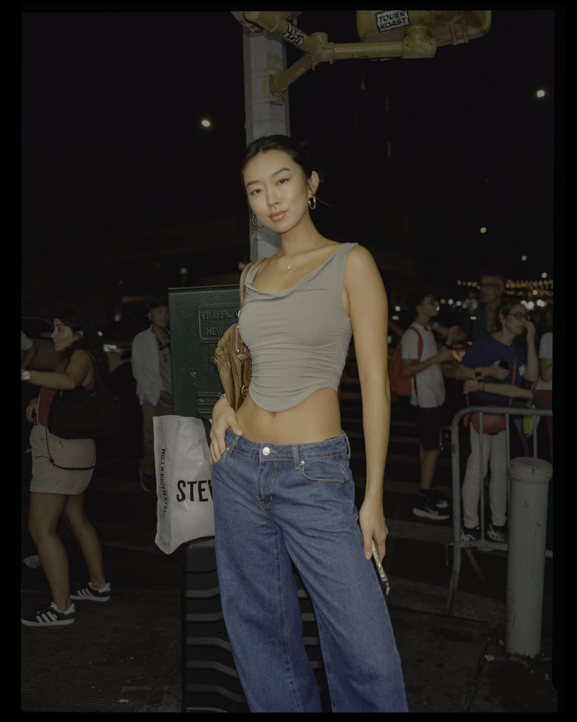 A young woman with long black hair, wearing a beige sleeveless top and baggy blue jeans, standing outdoors at night in a crowd with streetlights and buildings in the background.