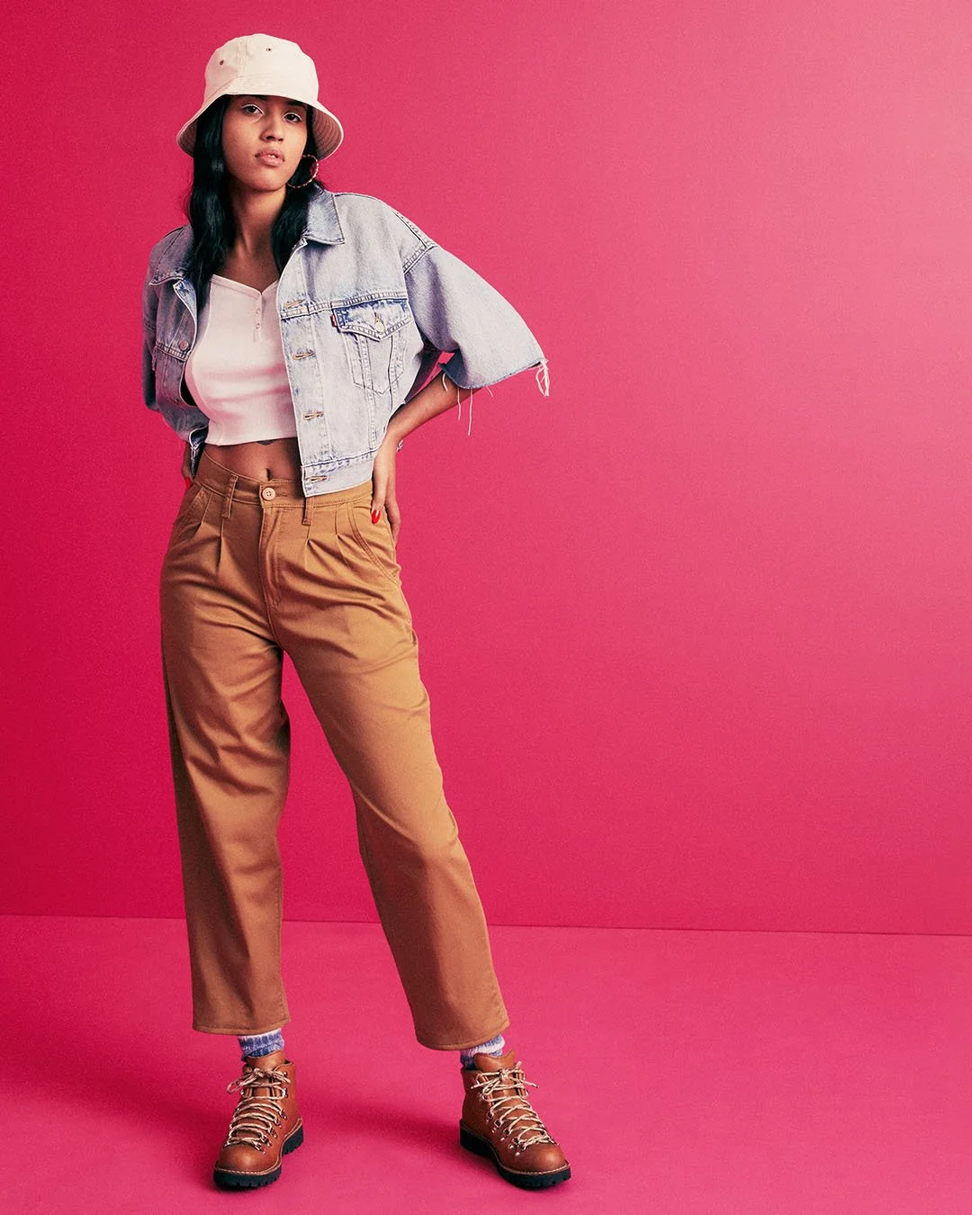 A woman wearing a beige bucket hat, denim jacket, crop top, tan pants, and tan boots standing against a pink background.