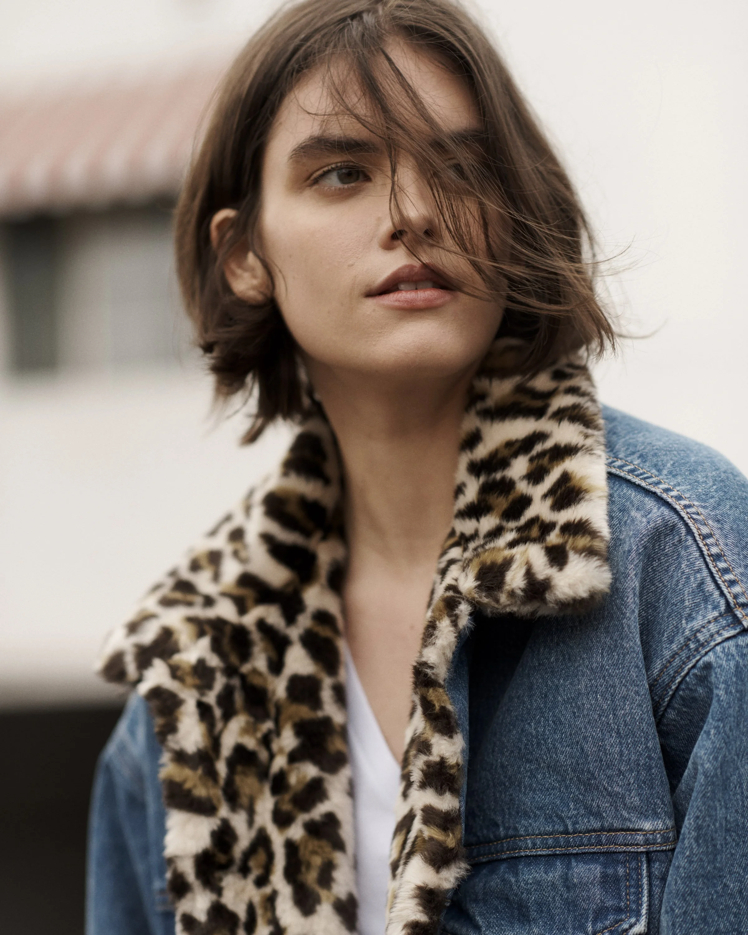 A young woman with short brown hair looking at the camera, wearing a leopard print scarf, a denim jacket, and a white shirt.