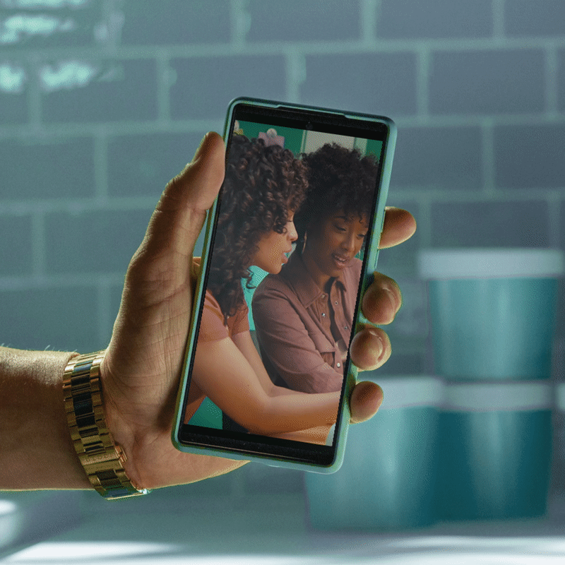 A person holding a smartphone with a photo of two women smiling and looking at each other. The background features a gray brick wall and a teal container.