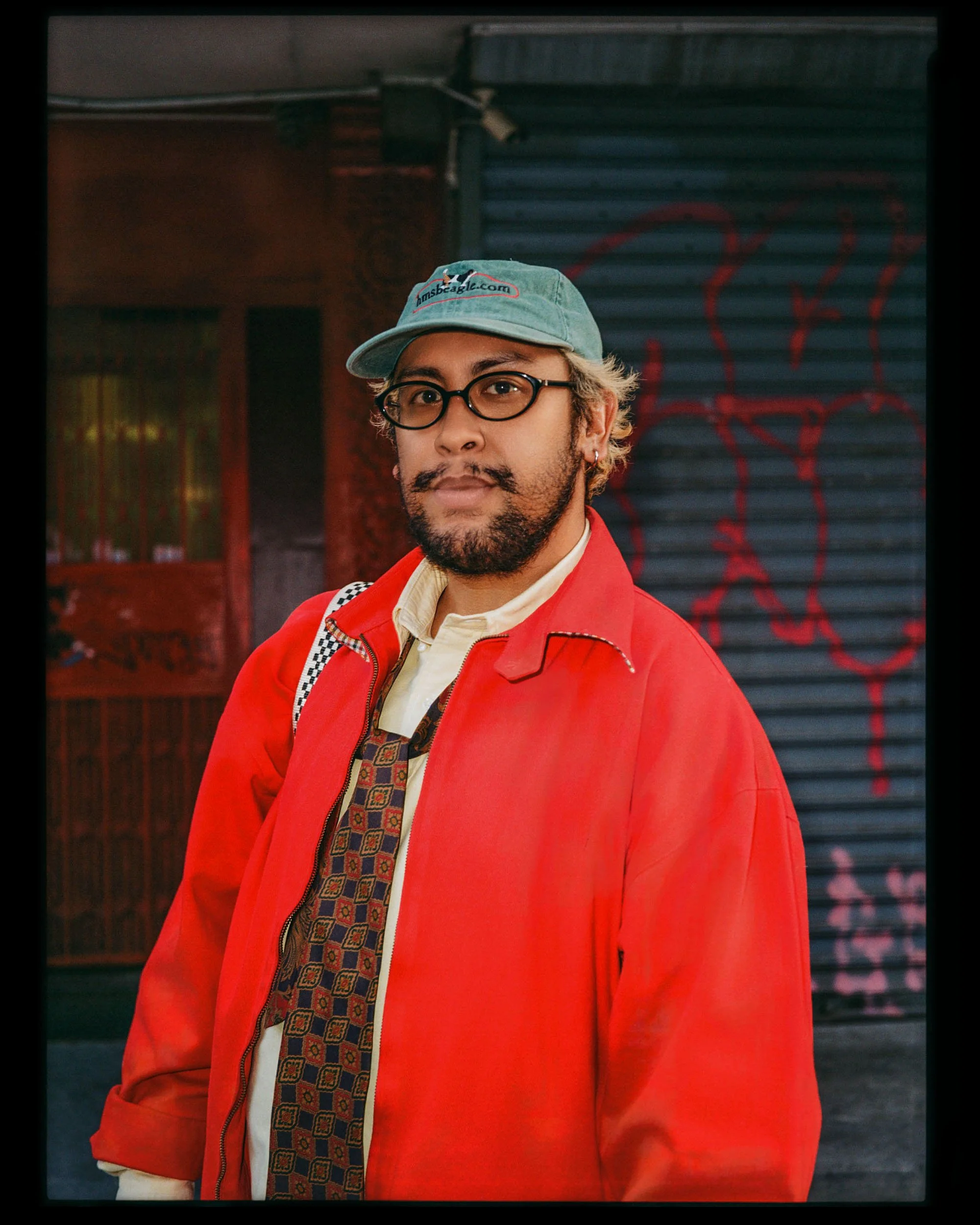 A man wearing glasses, a baseball cap, and a red jacket standing in front of a graffiti-covered wall.