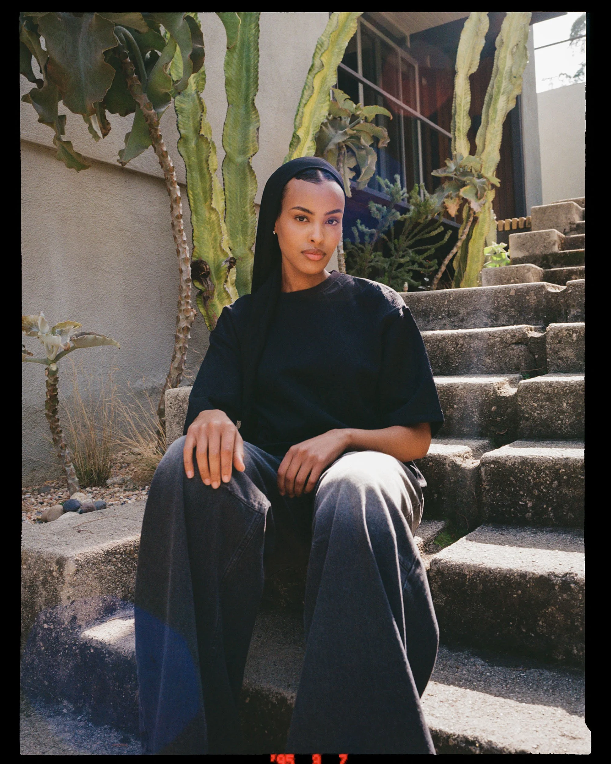 A woman with dark hair wearing a black shirt and gray pants, sitting on outdoor stone steps near tall cacti and desert plants, with a building in the background.