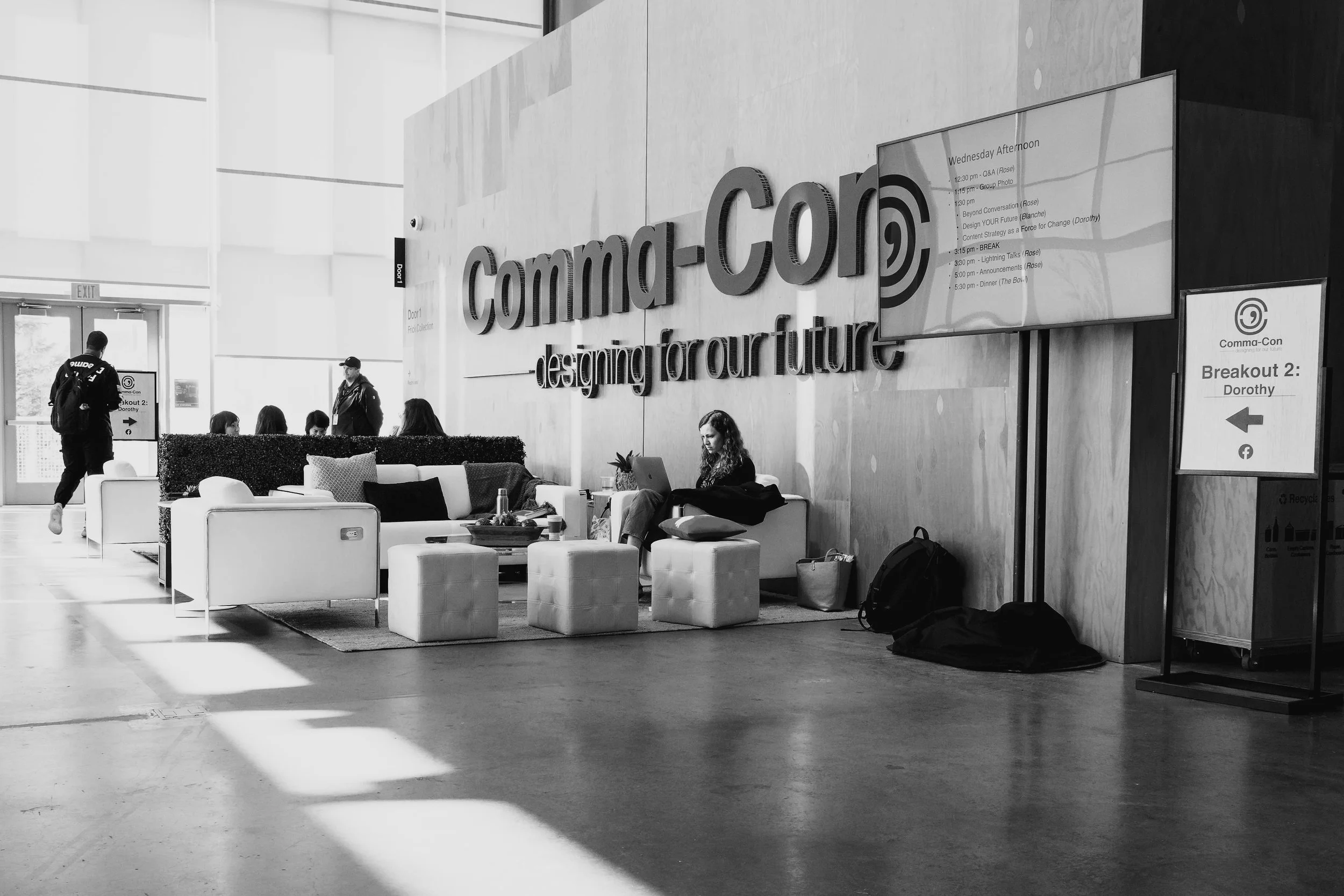 Interior of a modern conference or convention center with a large wall-mounted sign that reads "Comma-Con" and "designing for our future." There are people sitting on couches and chairs, some working on laptops, with a woman in the foreground focusing on her laptop. A sign indicating "Breakout 2: Dorothy" is visible, along with schedules and directional signs.