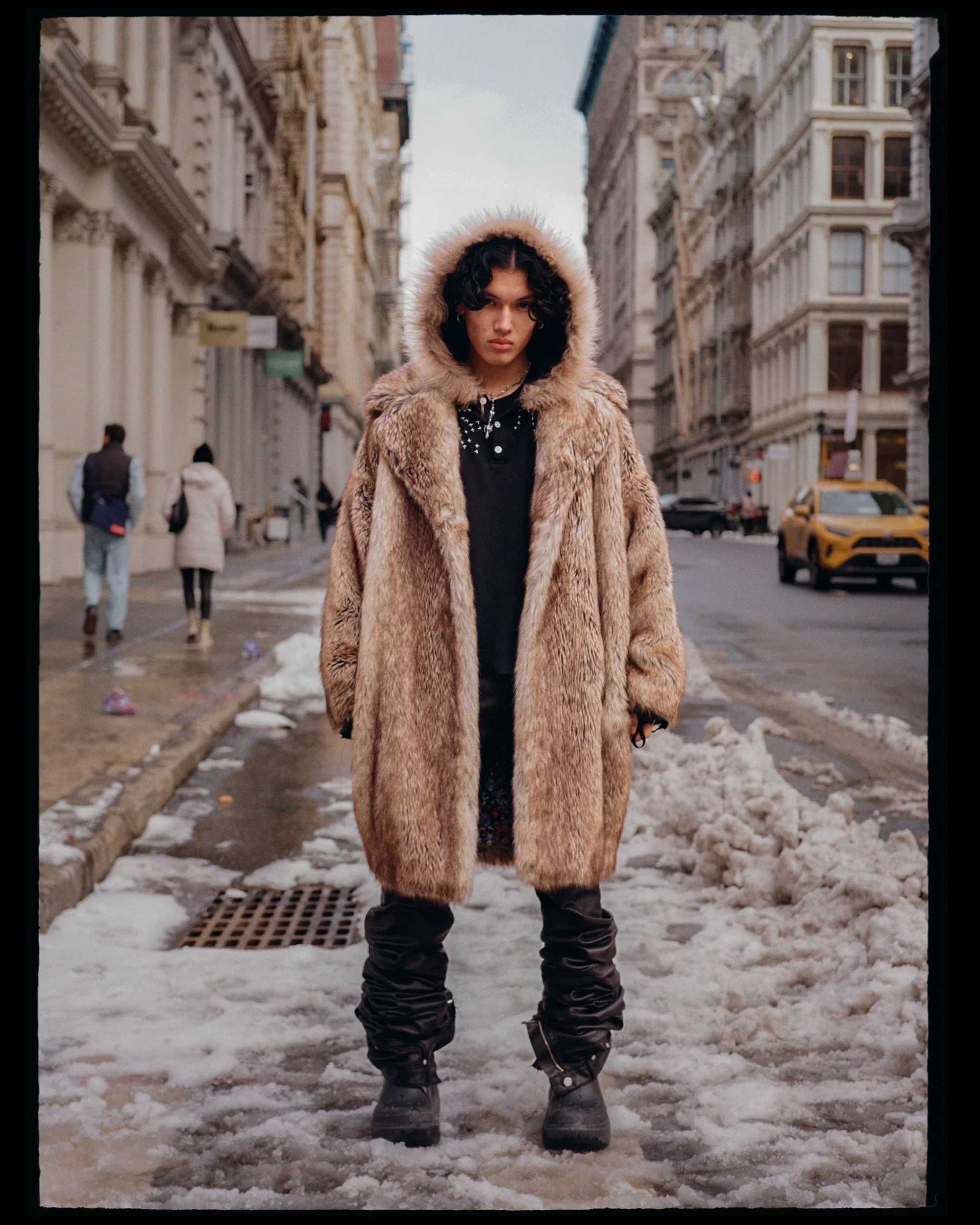 A person standing in the middle of a snow-covered city street, wearing a large fur coat with a hood lined with fur and dark winter boots. The street has some snow and slush, and there are buildings and people in the background.
