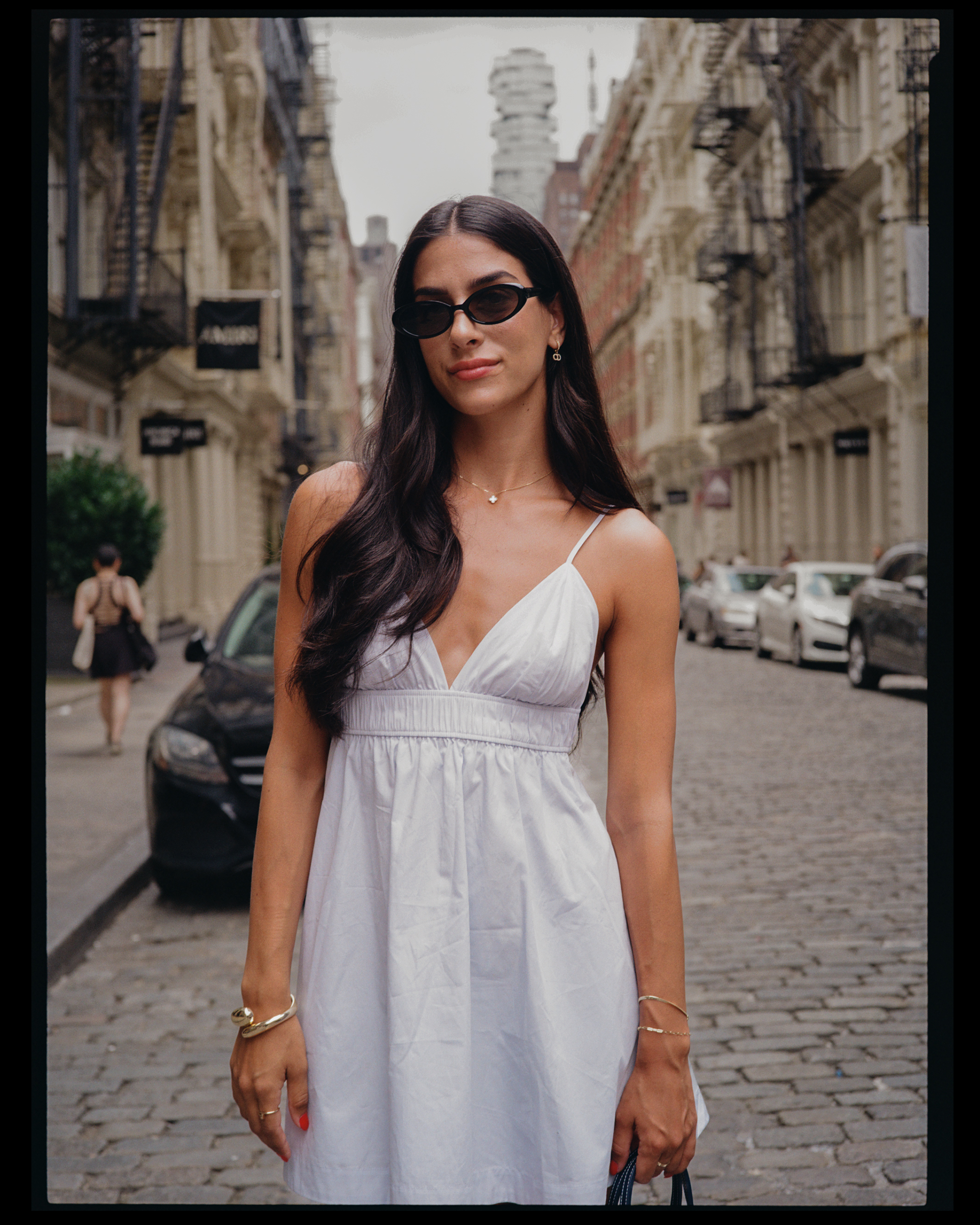 A woman with long dark hair wearing black sunglasses and a white dress standing on a city street with old buildings, parked cars, and pedestrians in the background.