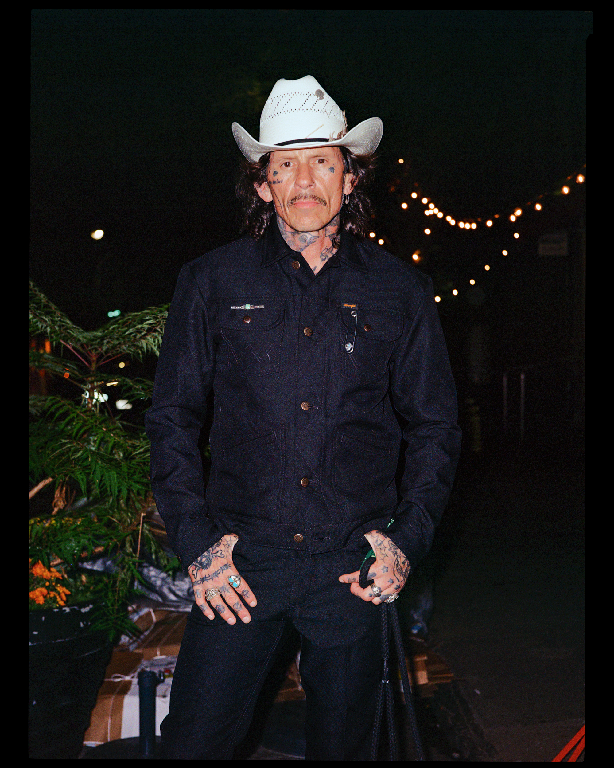 A man with tattoos and facial piercings wearing a white cowboy hat and dark denim shirt standing outdoors at night with string lights in the background.