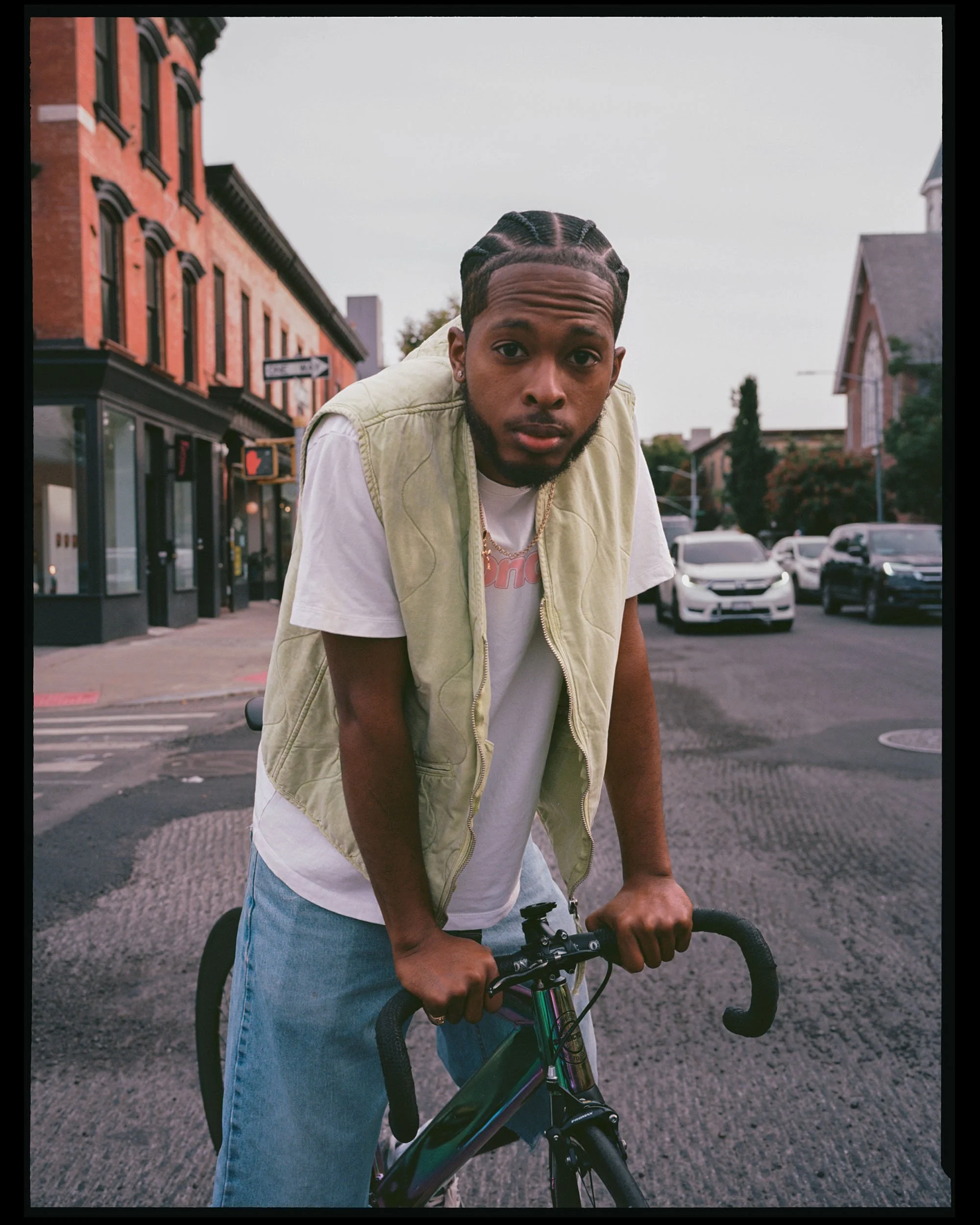 A young man with braided hair, a beard, wearing a yellow vest over a white t-shirt, riding a bicycle on an urban street lined with parked cars and brick buildings.
