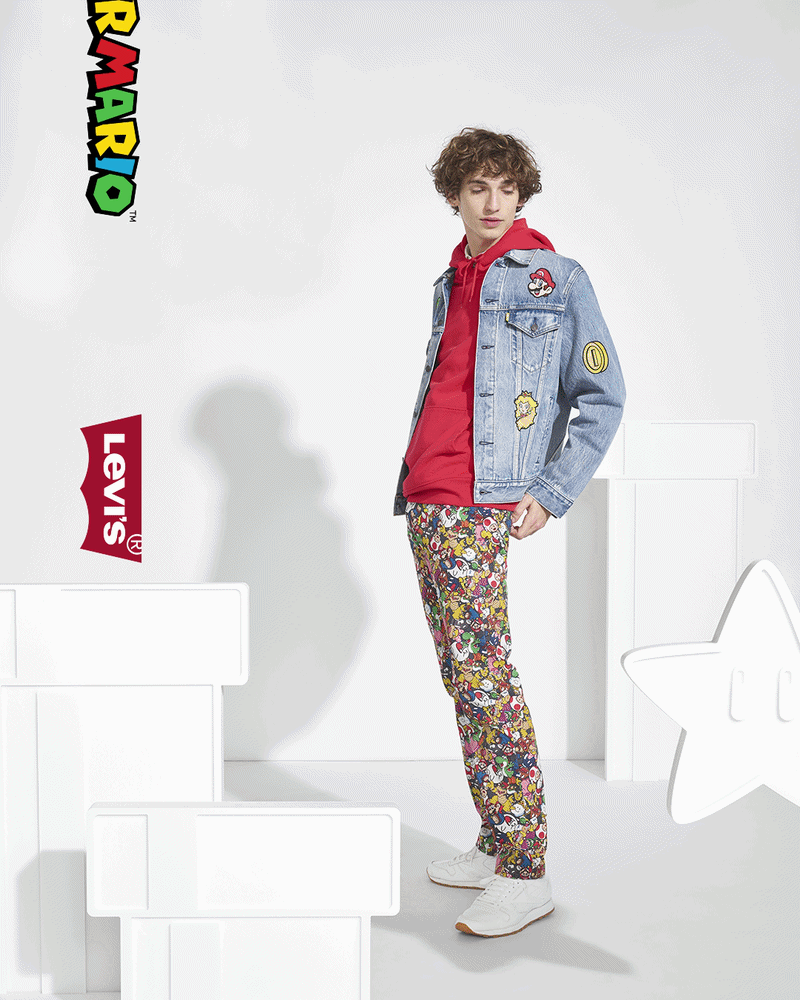 Young man with curly hair wearing a denim jacket with patches, a red hoodie, colorful patterned pants, and white sneakers standing in front of a plain background with LEGO and Levi's logos.