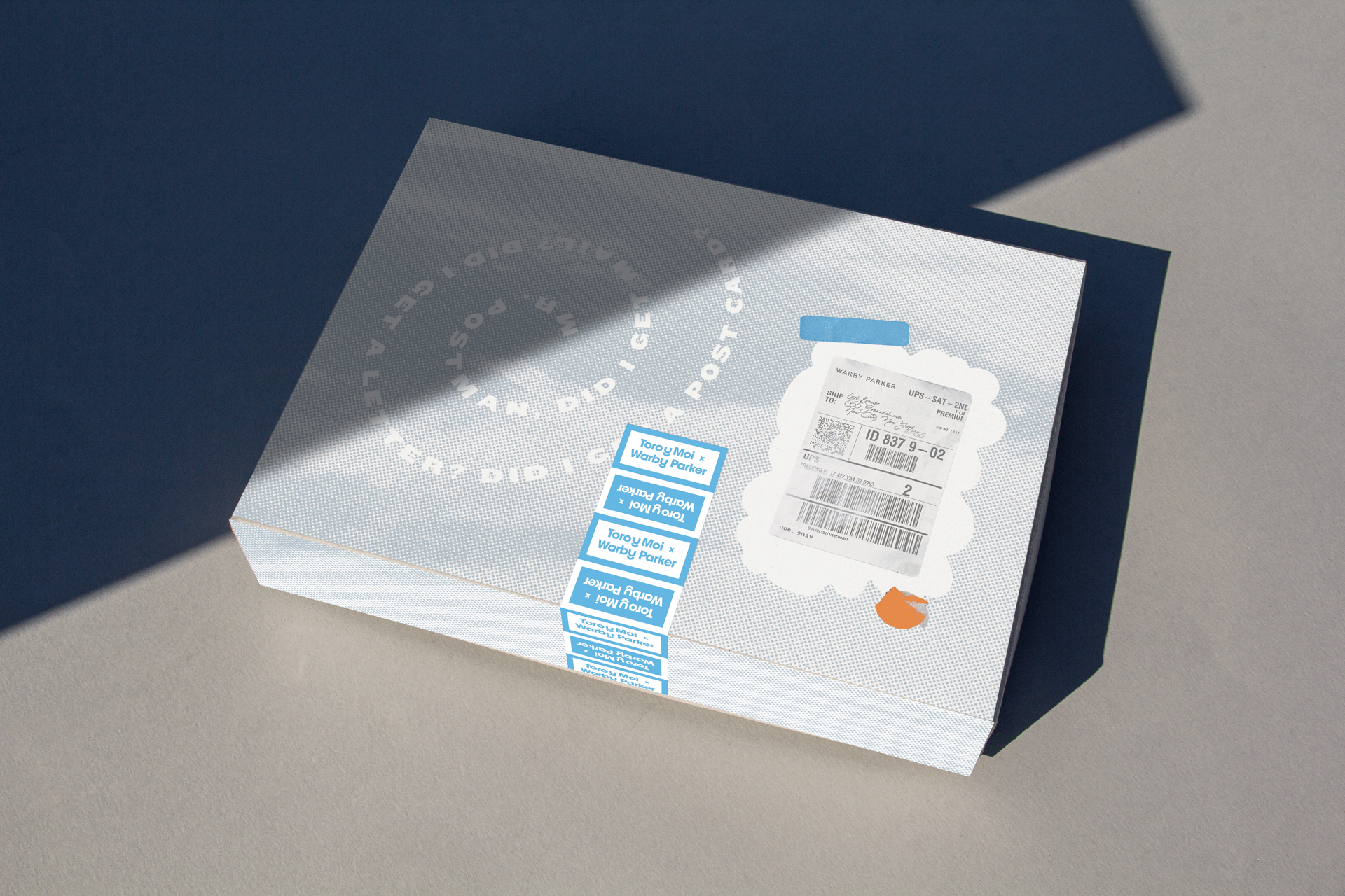White box with a cloud-shaped shipping label on top, accompanied by multiple blue and white stickers and a parcel label with tracking information. The box casts a shadow on a light gray surface.