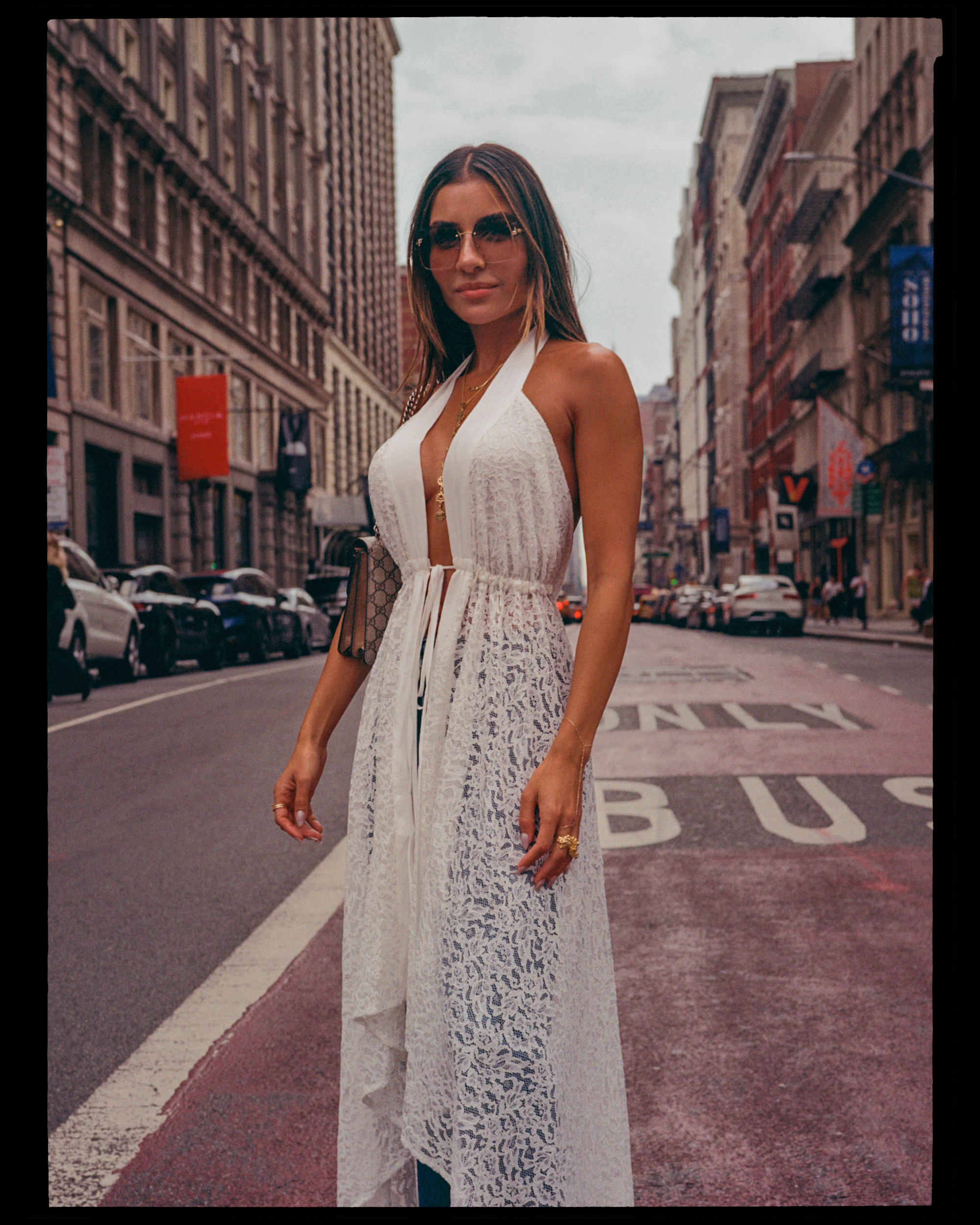A woman with long hair wearing sunglasses and a white lace dress standing on a city street with tall buildings and parked cars in the background.
