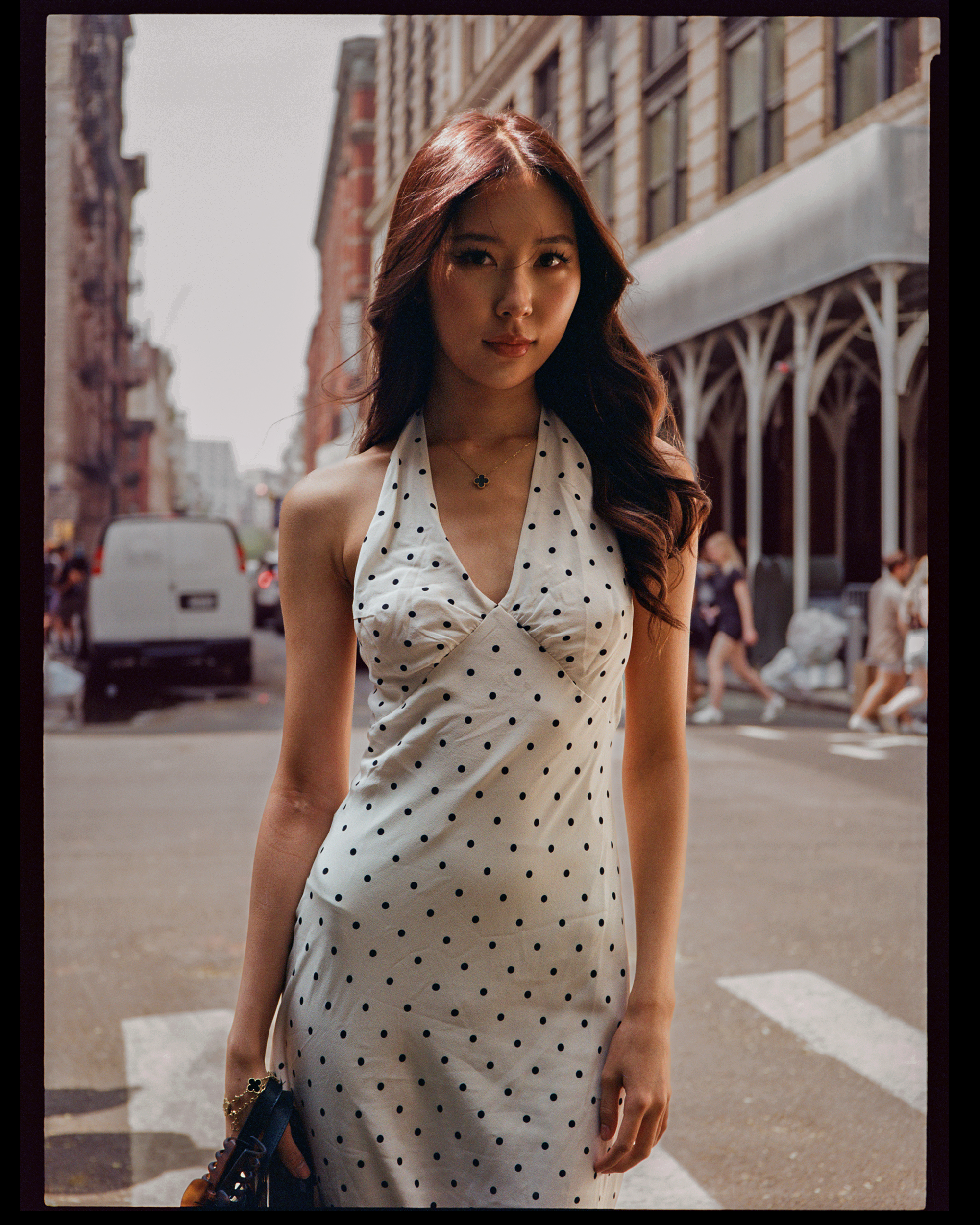 A young woman with long, wavy, reddish-brown hair standing on a city street, wearing a white dress with black polka dots and a necklace with a four-leaf clover pendant, holding a black purse with brown accents. The background includes buildings, peop