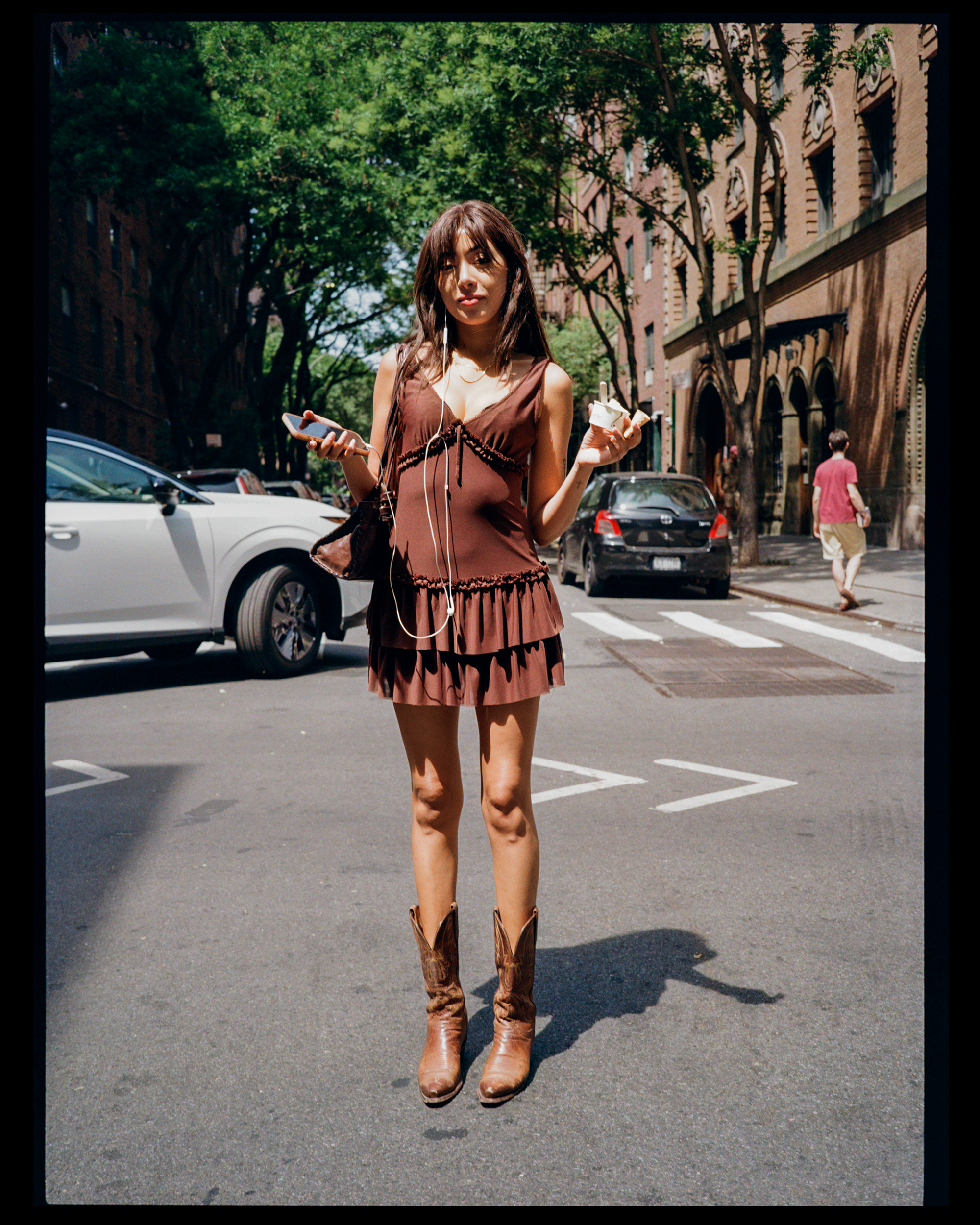 A woman in a brown dress and cowboy boots standing on a city street, holding a smartphone and an ice cream cone, with parked cars and brick buildings in the background.