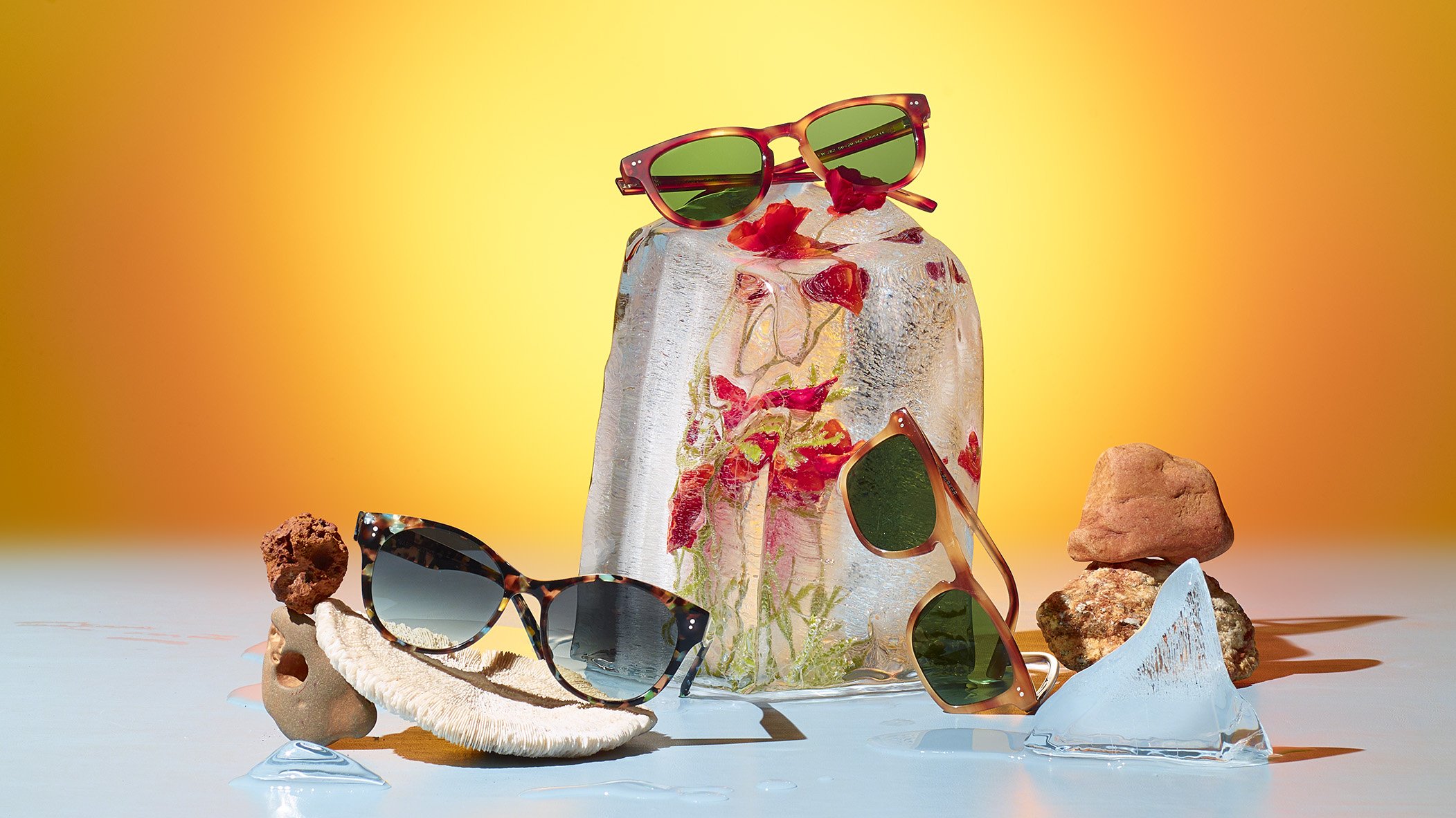Multiple pairs of sunglasses positioned on irregular objects and a glass block, with rocks and sea shells surrounding them, against a yellow-orange background.