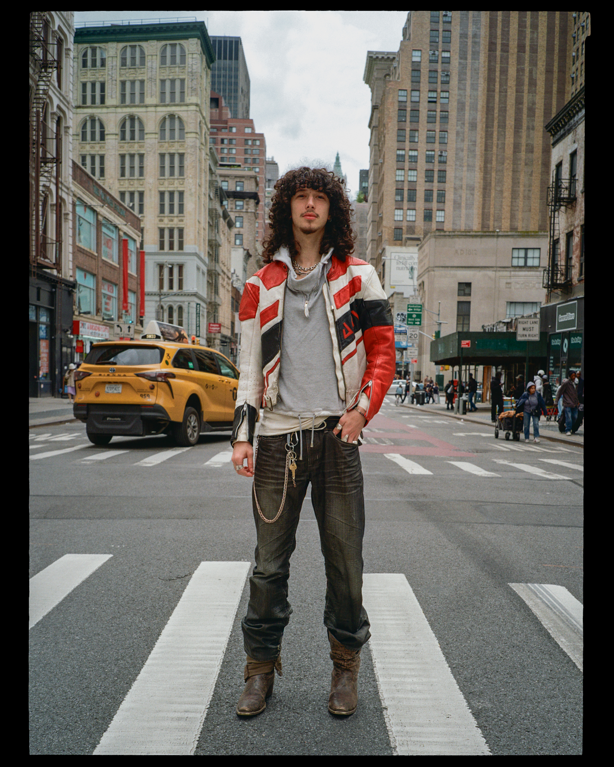 A young man with curly hair and a light goatee standing in the middle of a crosswalk in a city, wearing a red, white, and black jacket, gray hoodie, dark pants, and brown boots, with skyscrapers and city life around him.