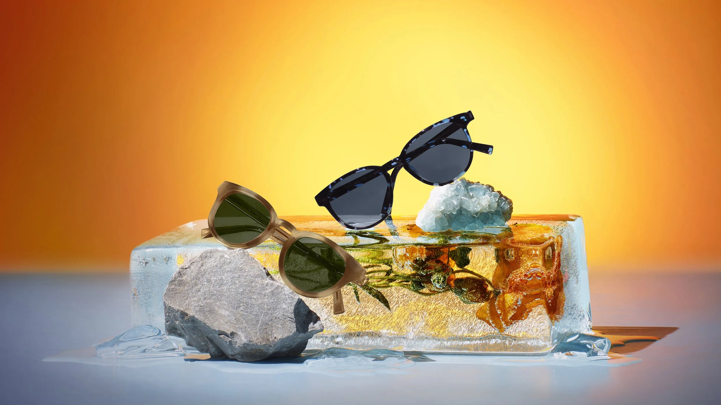 Two pairs of sunglasses, one black with blue accents and one beige, are placed on top of a clear ice-like block with rocks and decorative elements. The background is a gradient from orange to yellow.