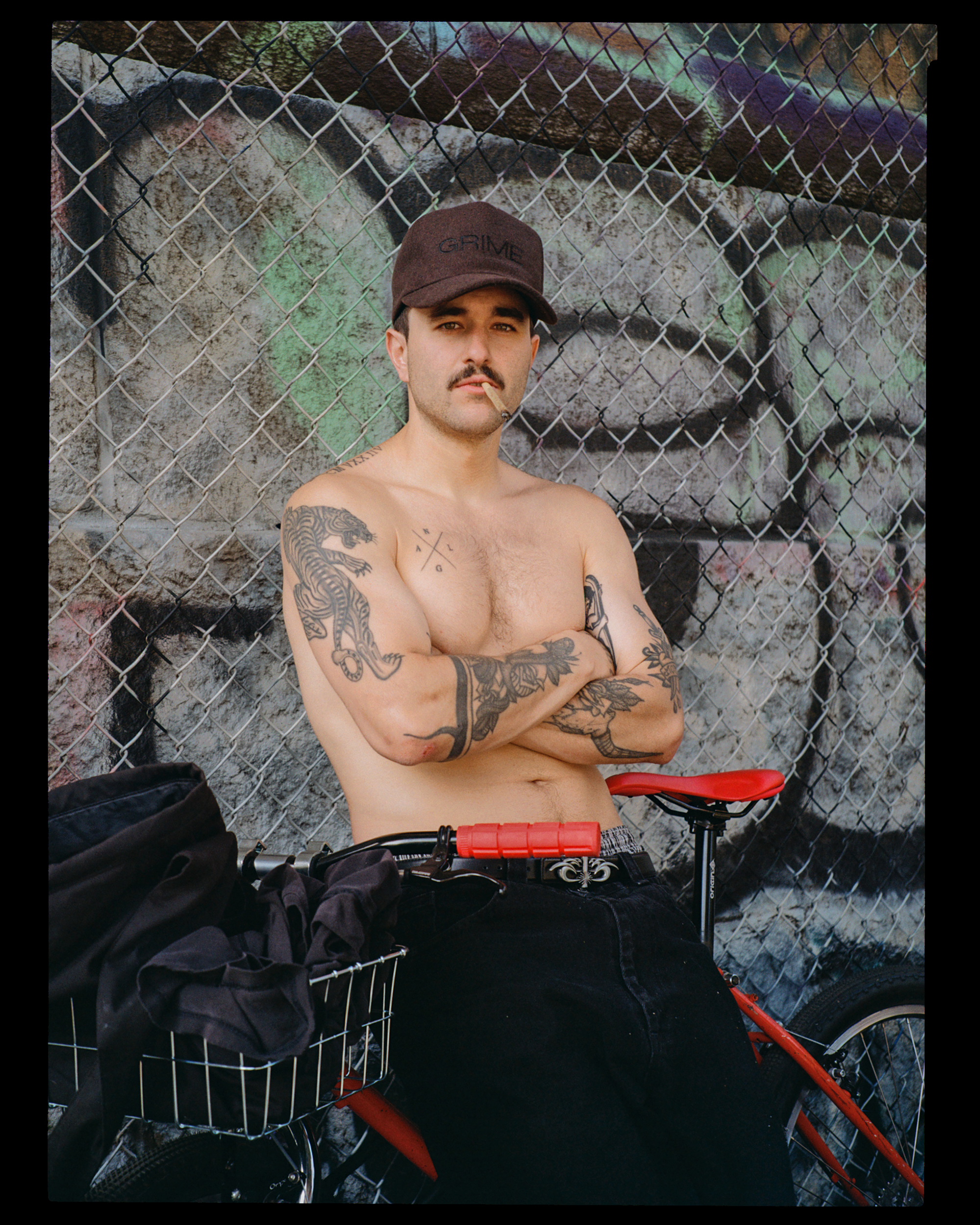 A shirtless man with tattoos, a mustache, and a cap that says 'GRIME' stands with crossed arms in front of a graffiti-covered brick wall and chain-link fence, with a red bicycle and black bag nearby.