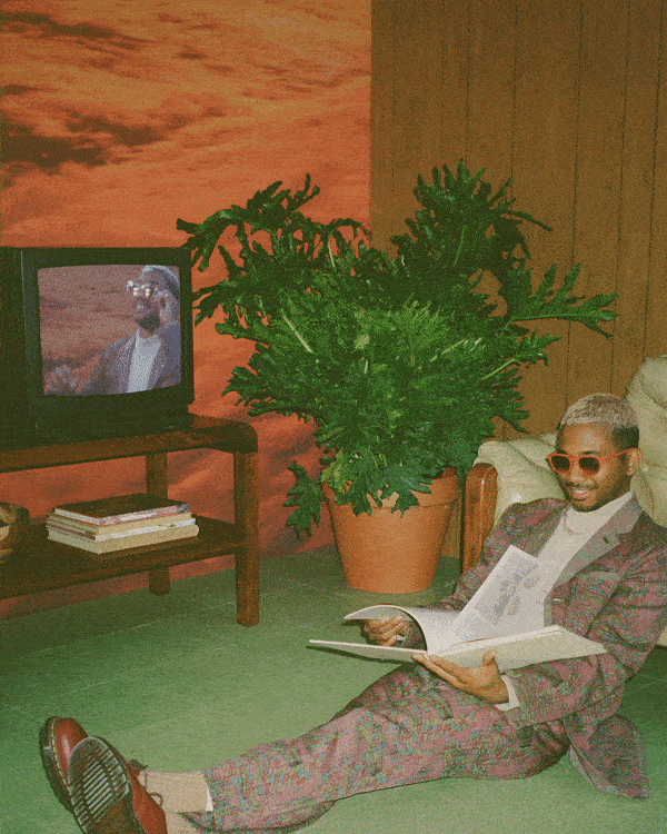 A man in a patterned suit and sunglasses sitting on the floor, leaning against a sofa, reading a magazine, with a TV displaying a sports scene and a large potted plant beside him.