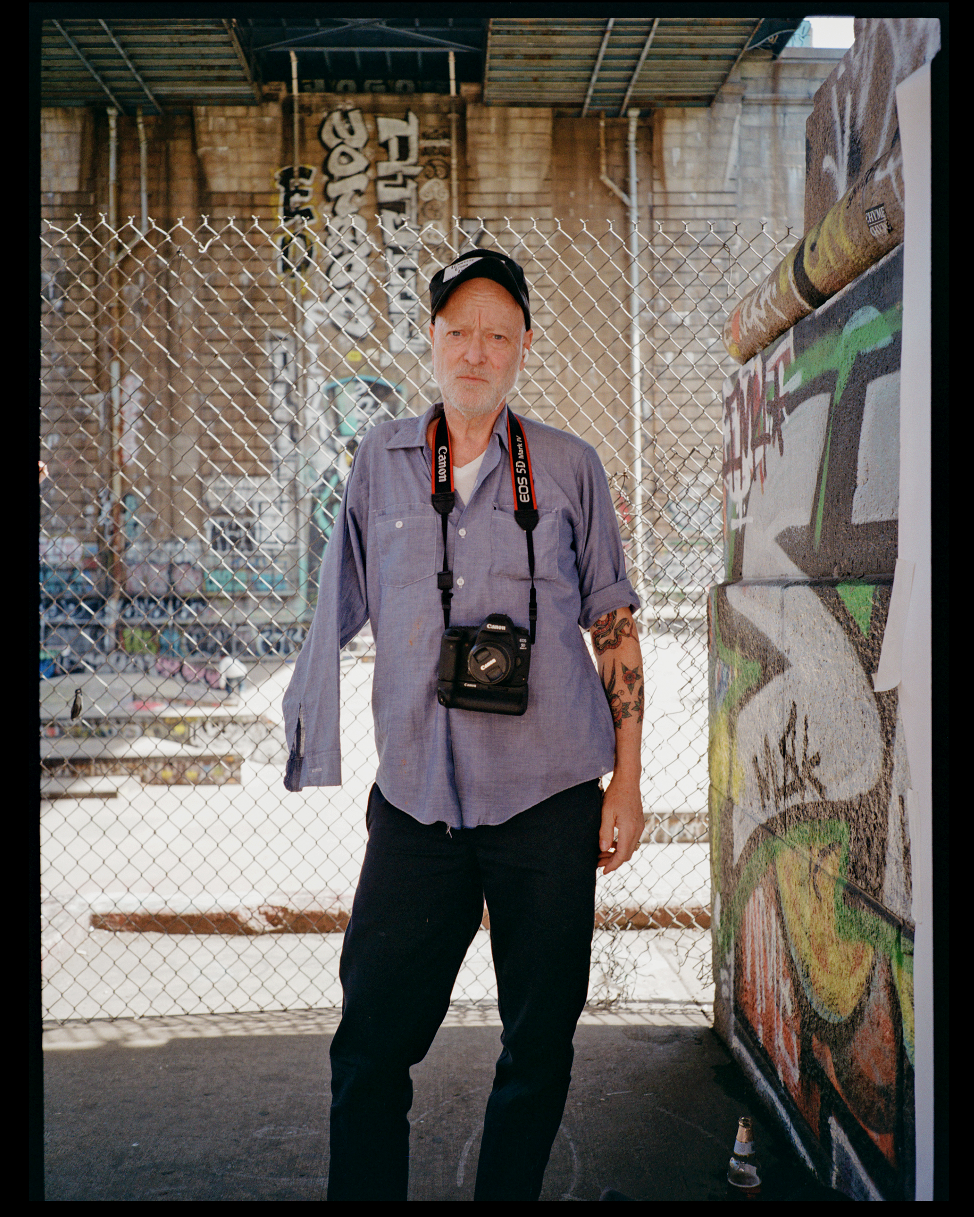 A man with a serious expression, wearing a black cap, gray shirt, and a camera around his neck, standing in front of a chain-link fence with graffiti art on the wall behind him.