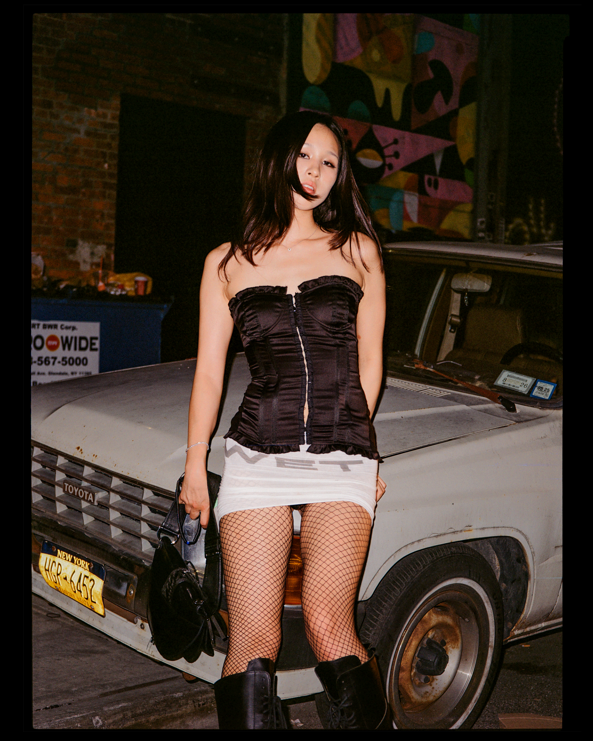 A young woman with light skin and dark hair, wearing a black strapless top, white skirt, fishnet stockings, and black boots, stands in front of an old, dirty white vehicle with a New York license plate. She is holding a black bag and standing at nigh