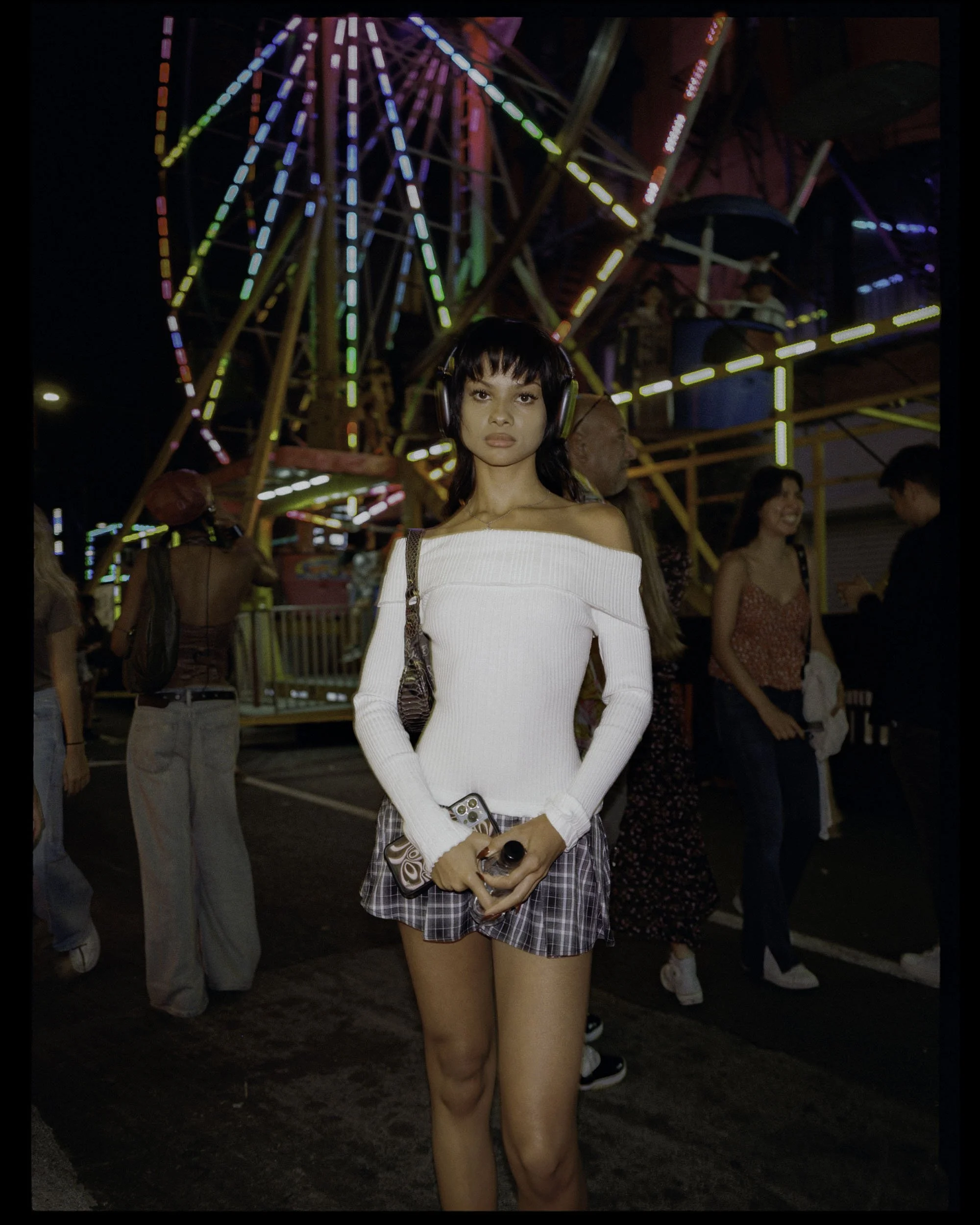 Young woman with short dark hair, wearing headphones, white off-shoulder sweater, and plaid skirt, standing at a nighttime amusement park with a Ferris wheel and other people in the background.