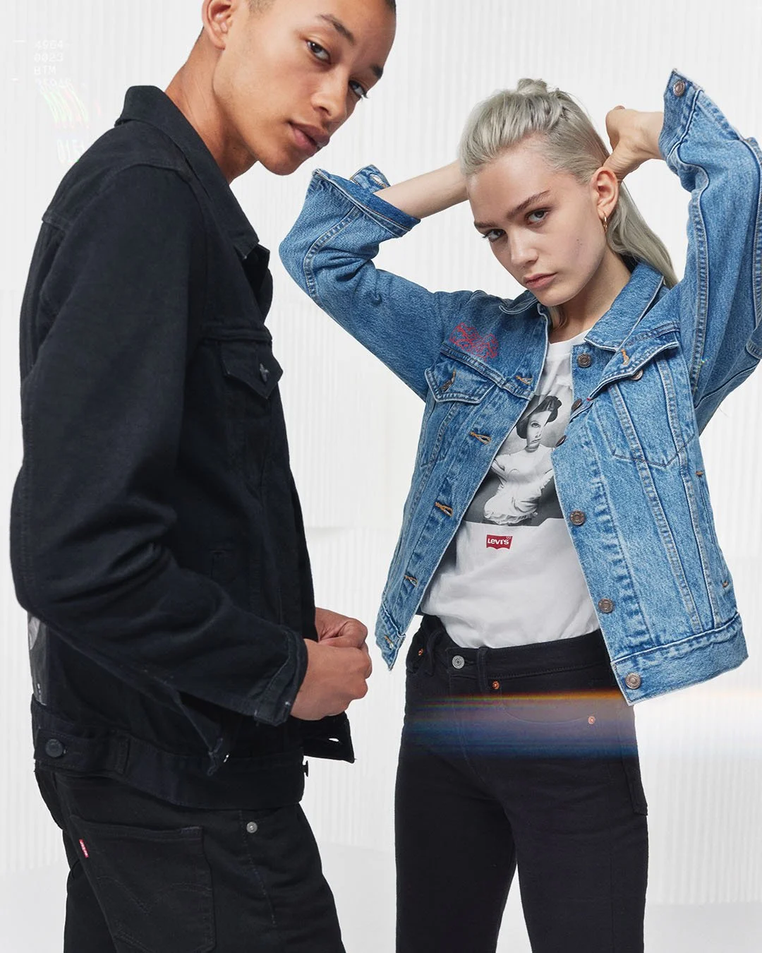 A young man and woman pose together in casual clothing. The man wears a black denim jacket and black pants, while the woman wears a blue denim jacket over a graphic T-shirt and black pants. They stand against a white background.