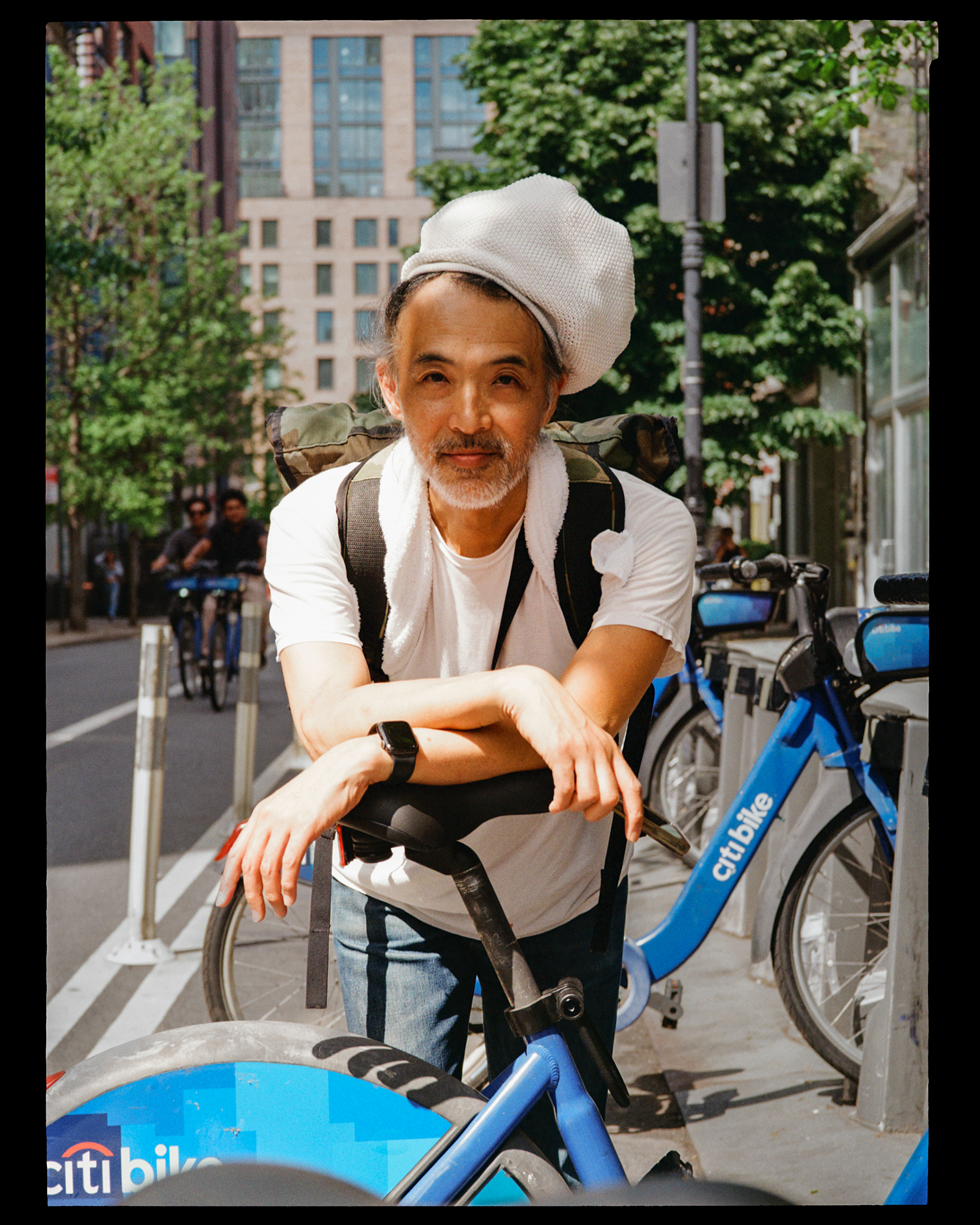A man with gray hair and a beard riding a blue Citi Bike, wearing a white shirt and a white towel around his neck, with a white hat on his head, on a city street with tall buildings and green trees in the background.