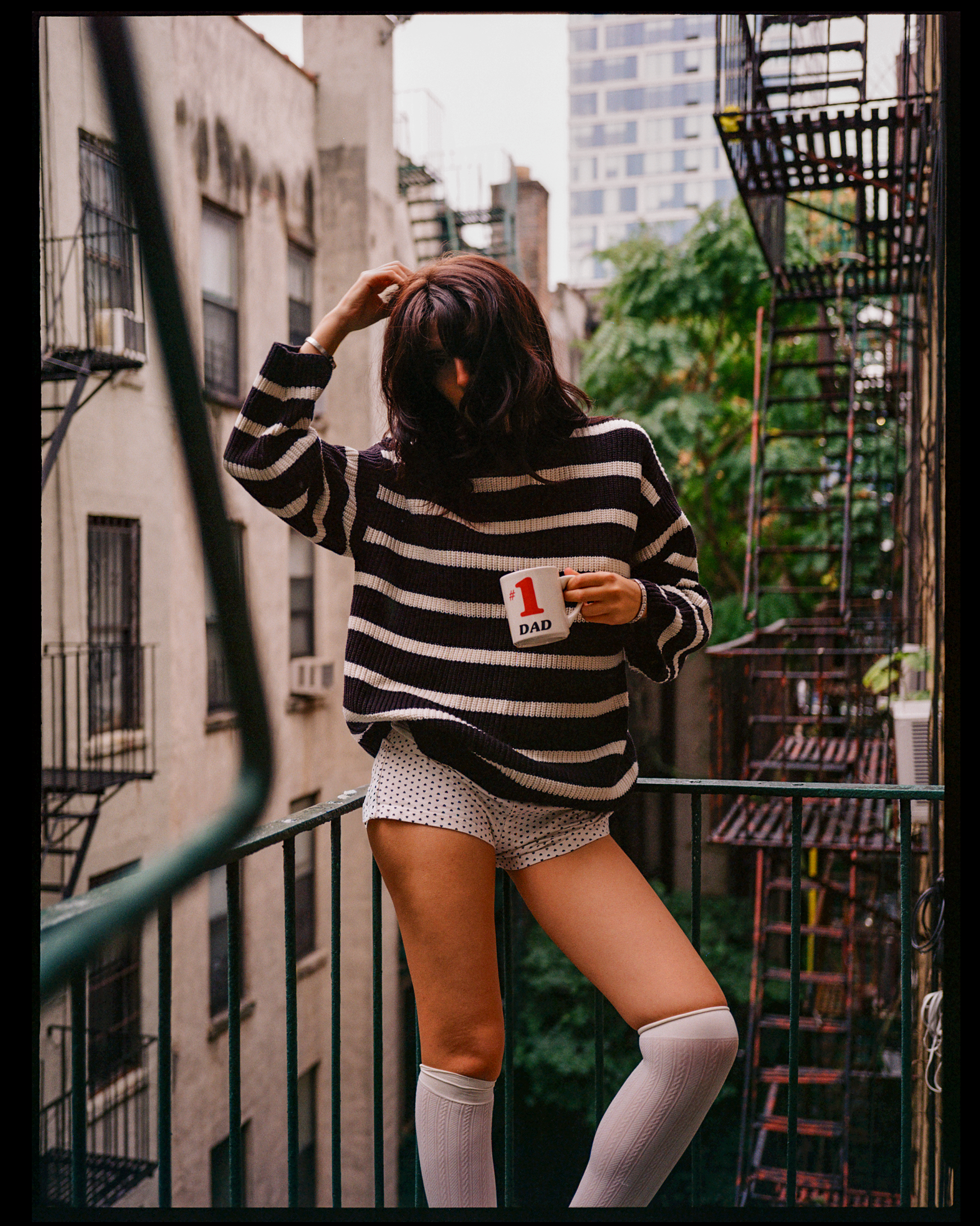 A woman standing on a fire escape balcony wearing a black and white striped sweater, white shorts, and thigh-high white socks with a pattern, holding a white mug that says '#1 Dad'. She is adjusting her hair with her right hand, with her face partial