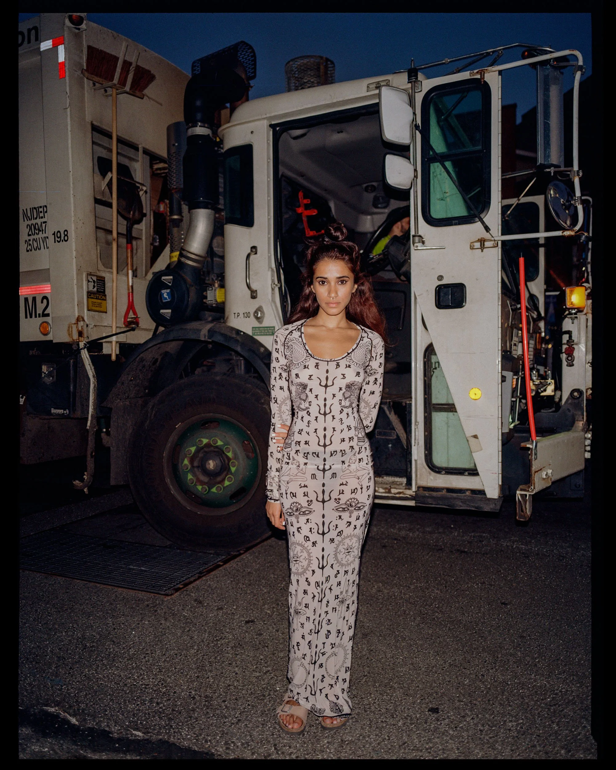 A young woman with long reddish-brown hair styled in a high bun, standing in front of a large utility truck at night. She is wearing a long white dress with black tattoo-style designs and beige open-toe sandals.