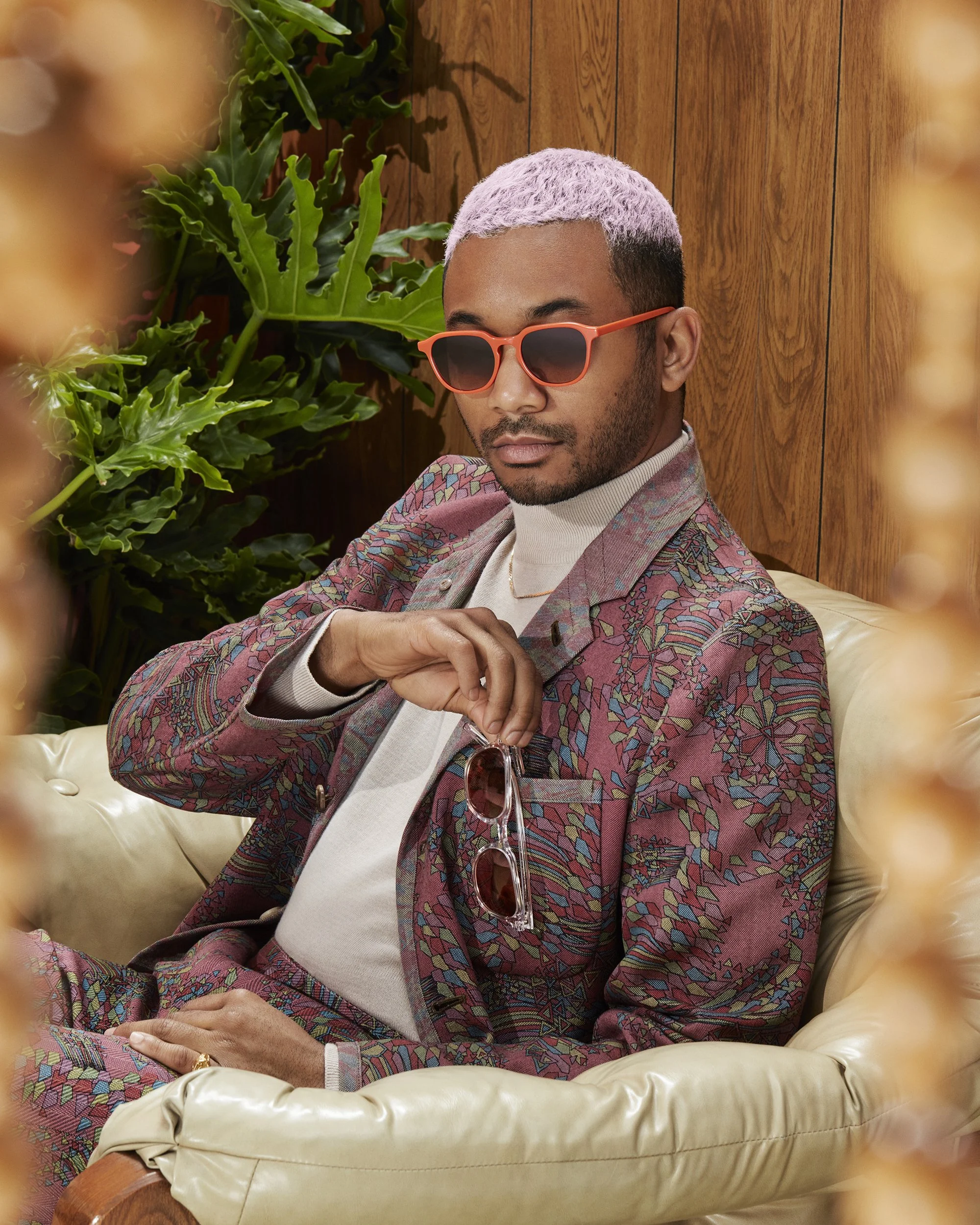 A man with light purple hair, wearing orange sunglasses, a patterned blazer, and a turtleneck sweater, sitting on a cream-colored tufted leather sofa with a wooden paneled wall and green plants in the background.