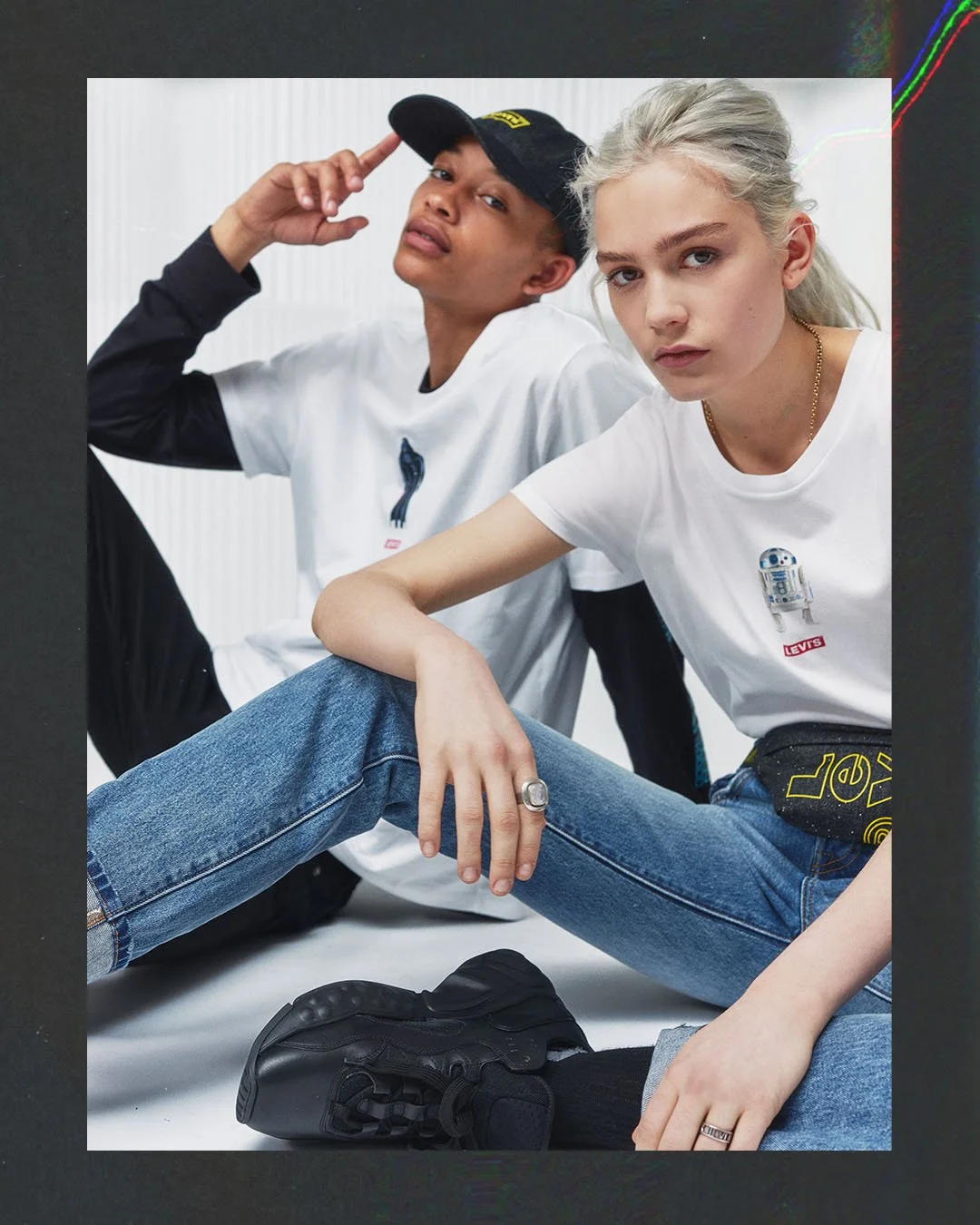 Two young models, one male and one female, sitting on the floor against a black and white background. They are wearing casual outfits with Levi's T-shirts and jeans, and looking at the camera with a serious expression.