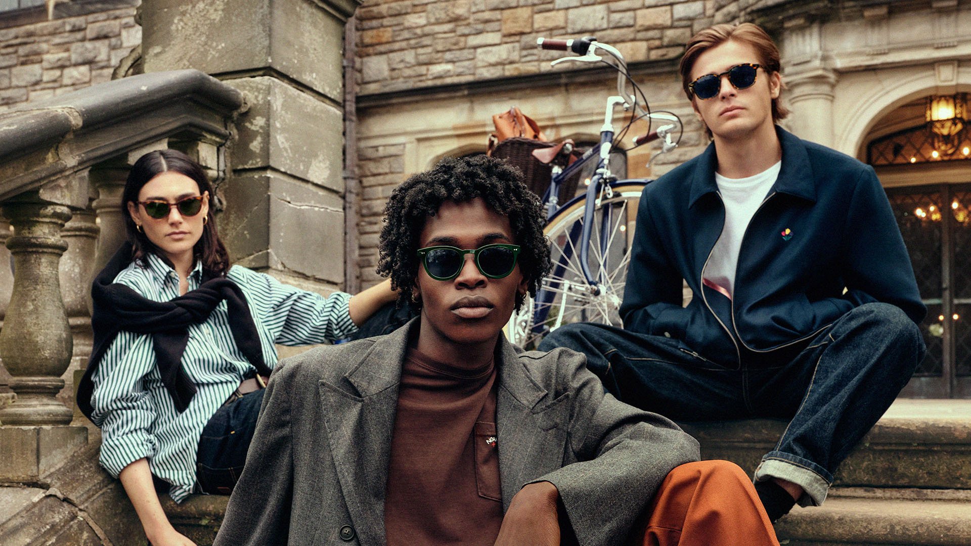 Three young adults sitting on stone steps outside a brick building, wearing sunglasses, with a bicycle in the background.