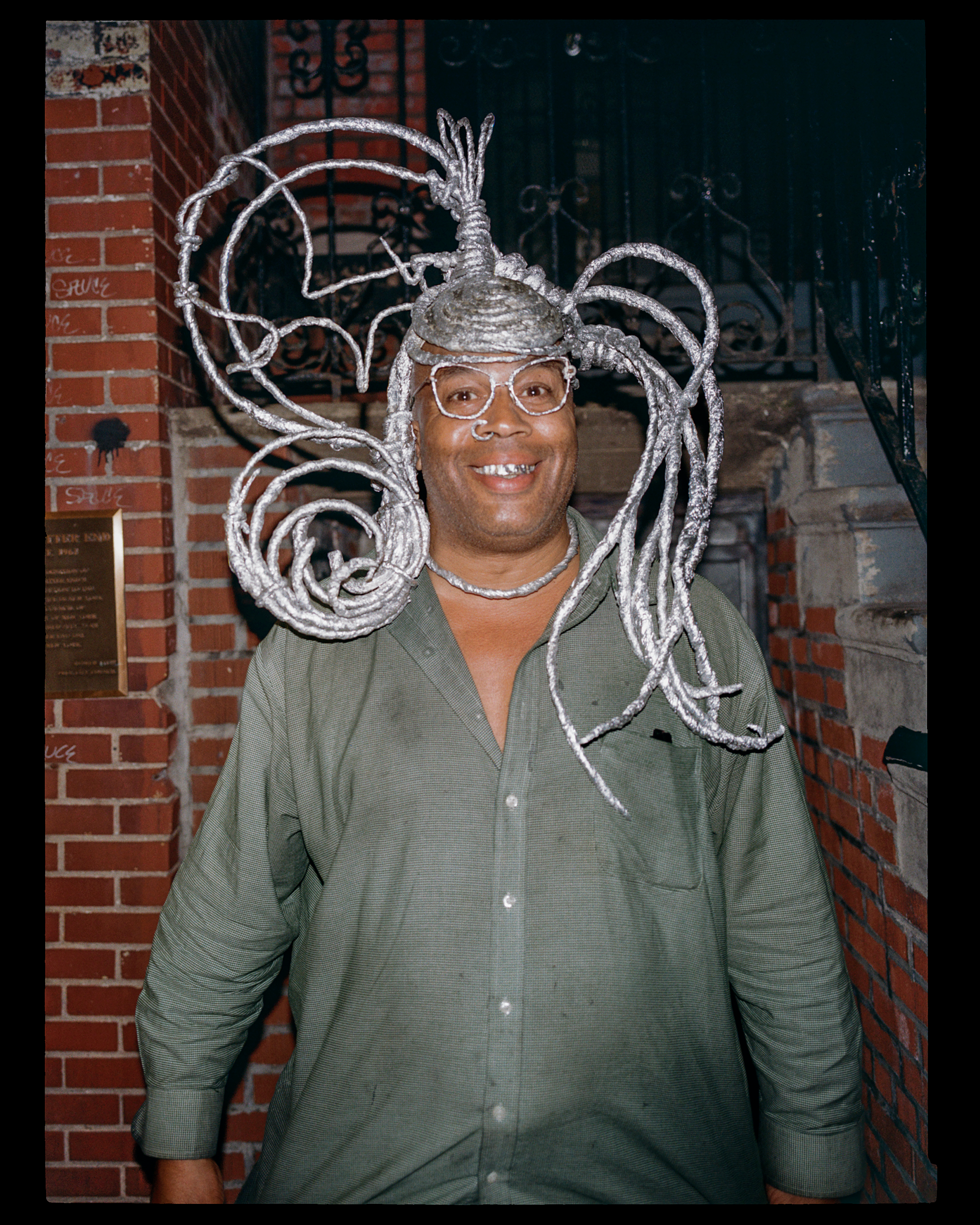 Person wearing a silver, artistic metallic headpiece with intricate loops and a crown-like structure, standing in front of a brick wall and black wrought iron gate, smiling.