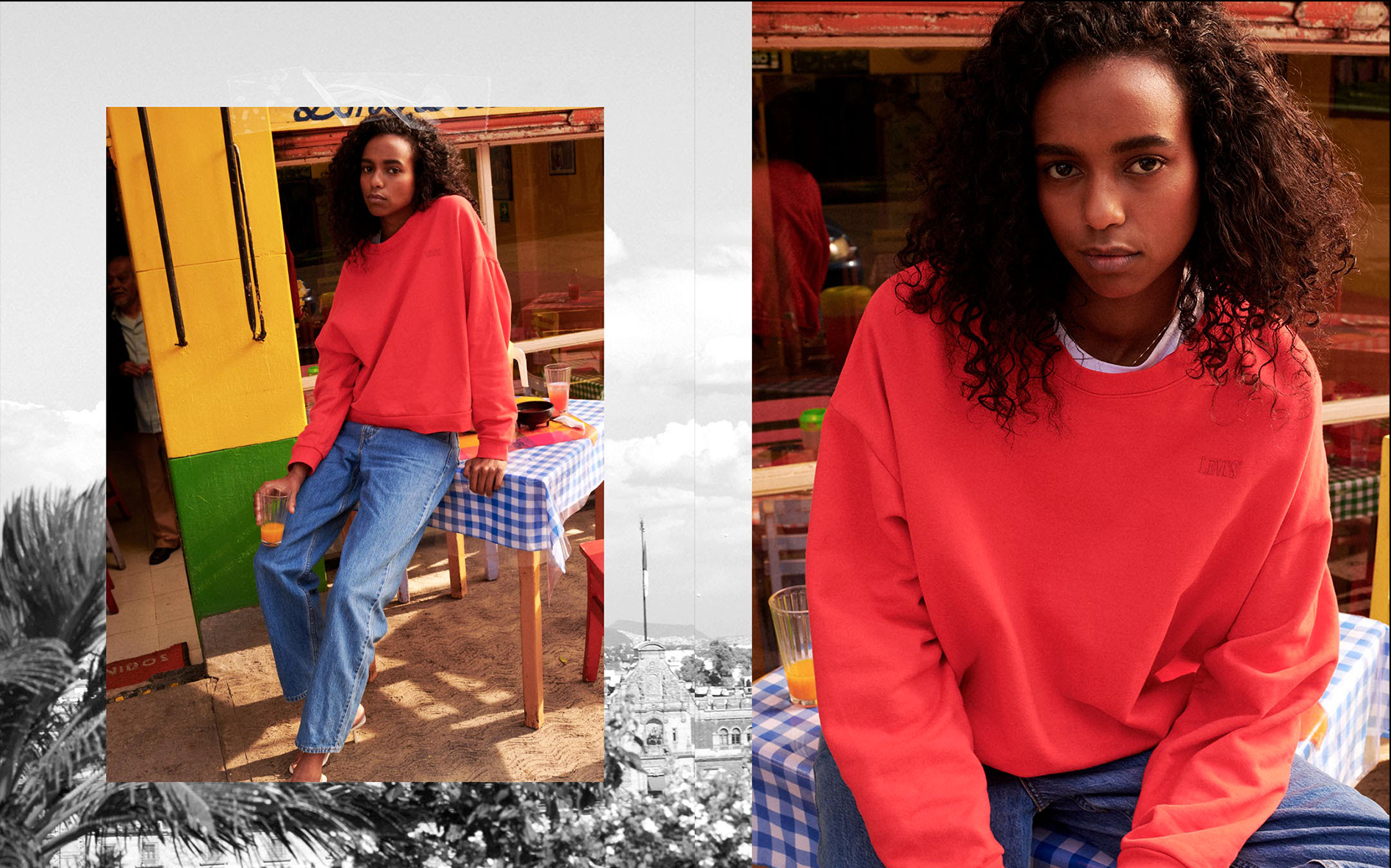 A young woman with curly black hair wearing a red sweatshirt and blue jeans, sitting at an outdoor restaurant or café with a checkered tablecloth, holding a glass with an orange drink, with a yellow and green painted wall behind her.