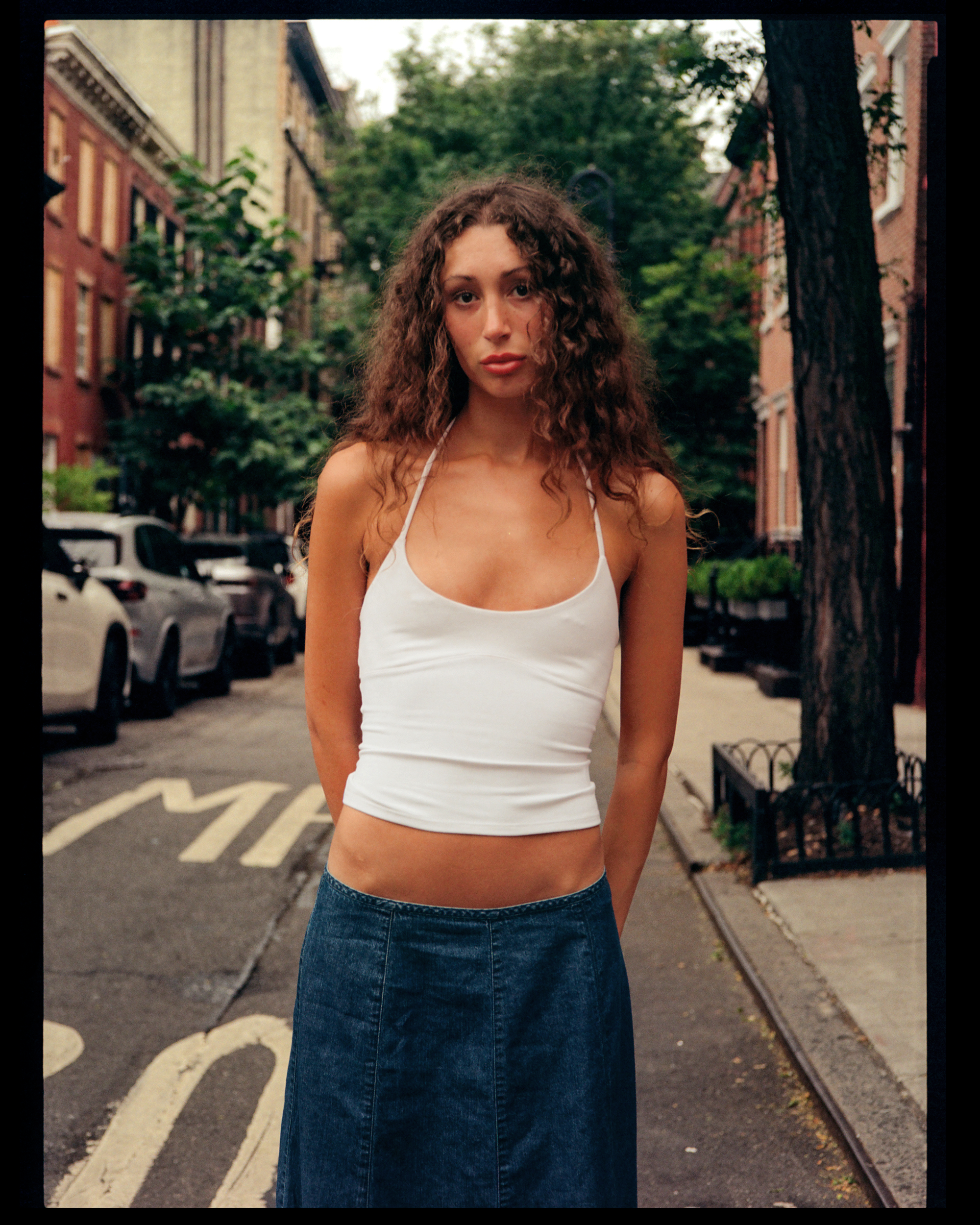 A woman with long curly hair wearing a white tank top and a denim skirt standing on a city street with parked cars, trees, and buildings in the background.