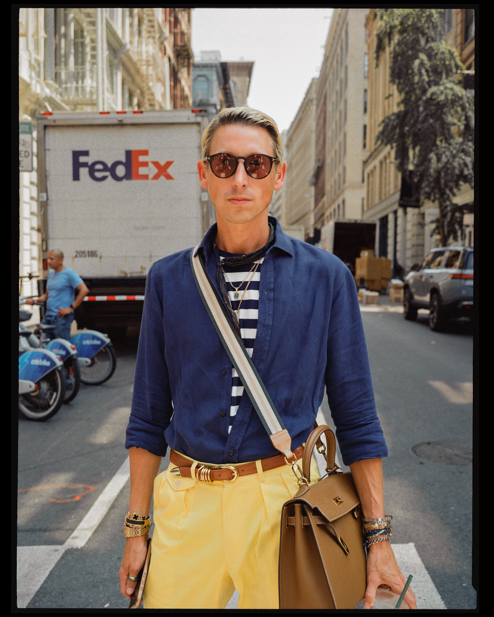 Person walking on city street wearing sunglasses, a blue shirt, yellow pants, and carrying a brown handbag.