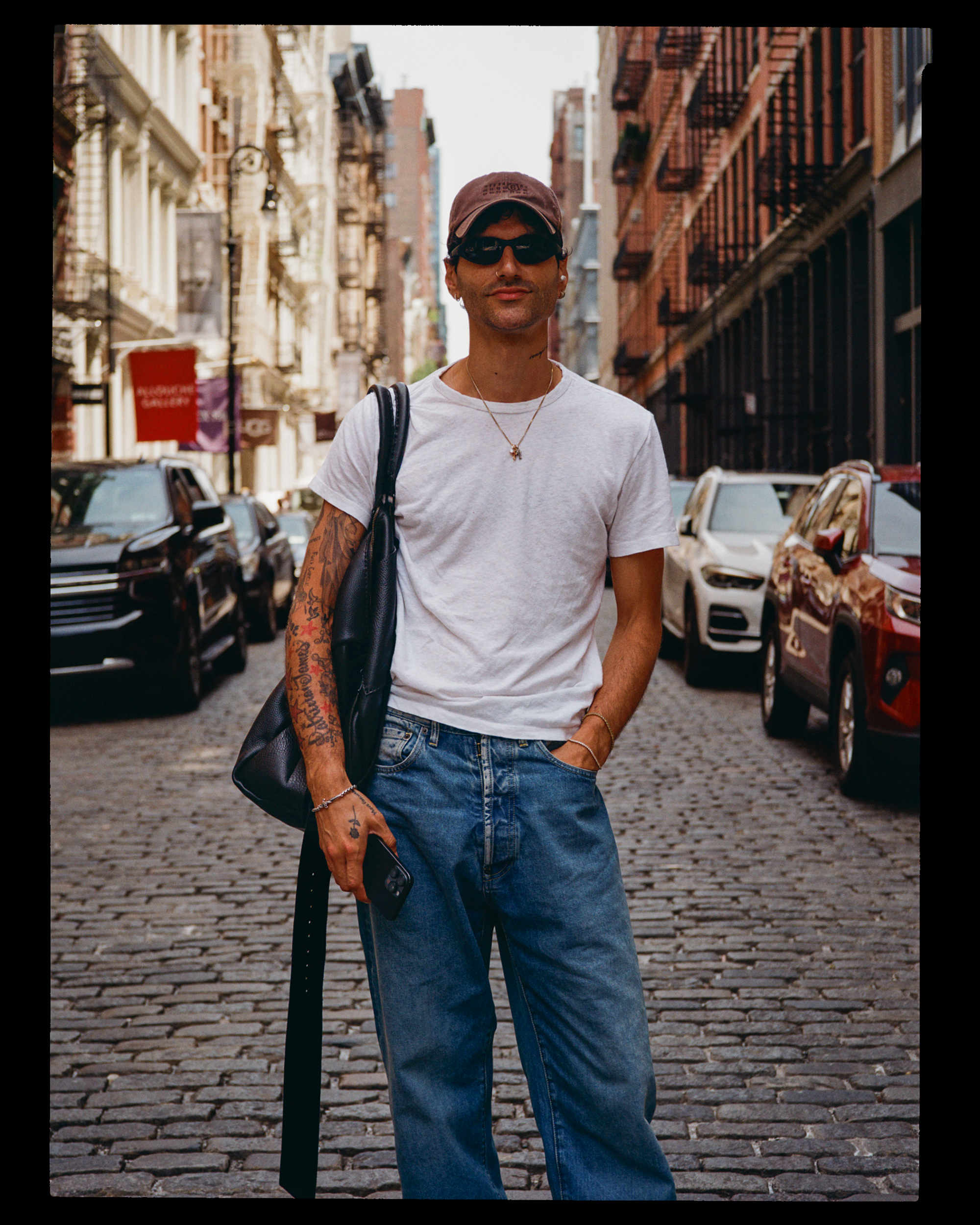 A person walking on a city street with brick buildings and parked cars, wearing sunglasses, a cap, a white T-shirt, and jeans, carrying a black bag and holding a phone.