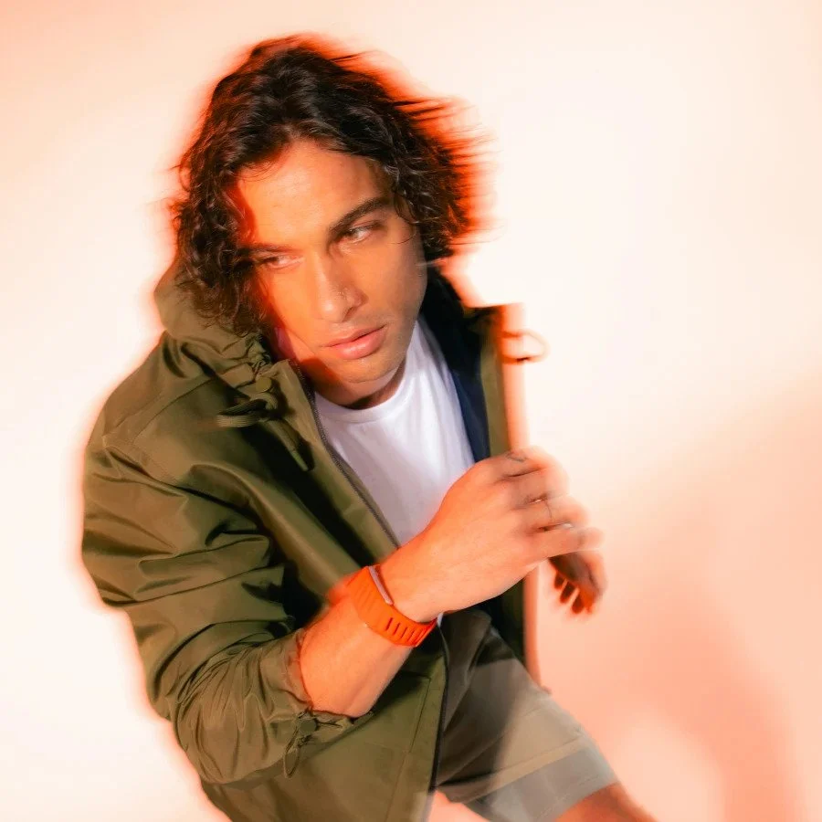 A man with curly dark hair and a serious expression, wearing a green jacket and white t-shirt, holding a wooden stick, with a red-orange glow and motion blur in the background.