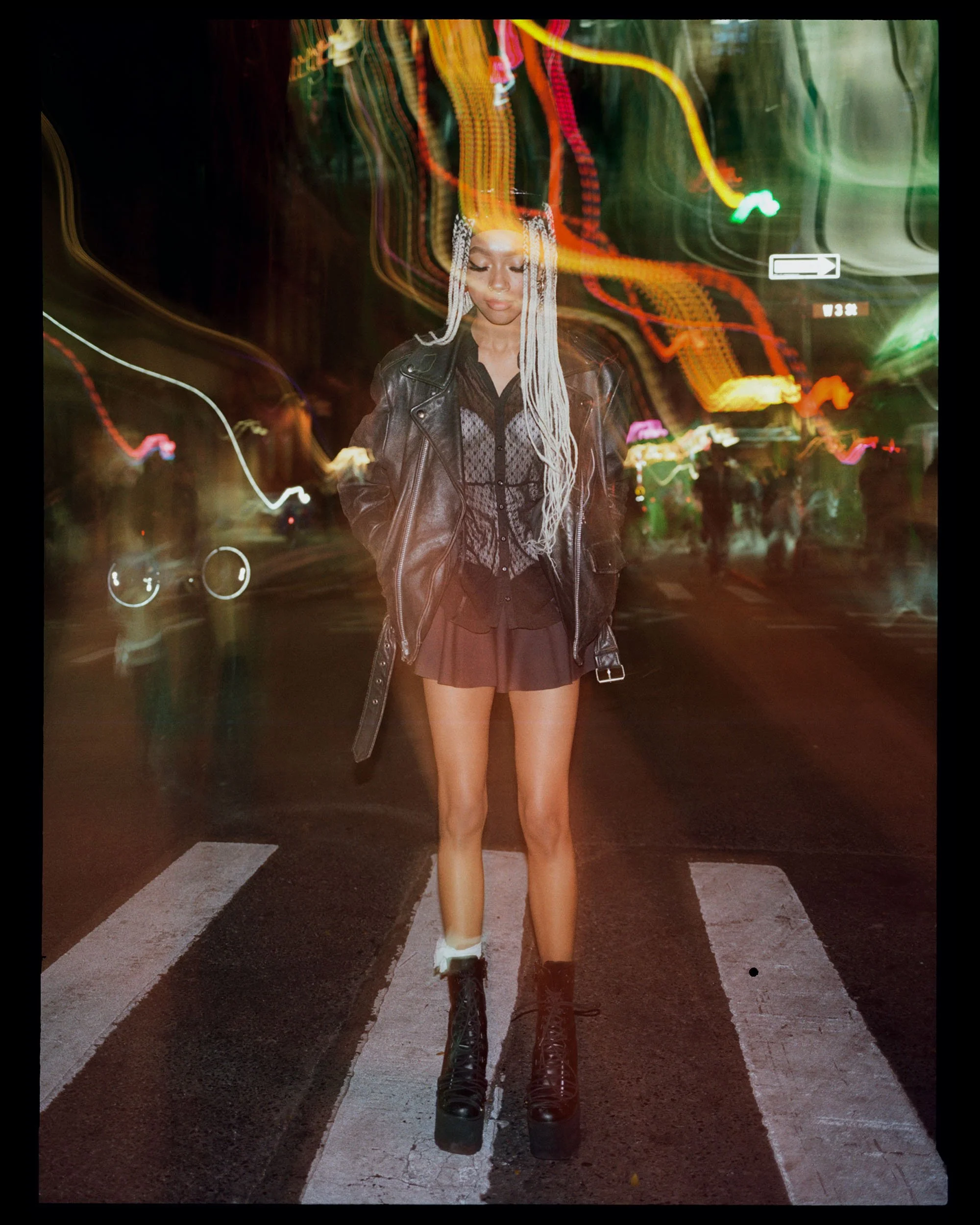 Young woman standing on a crosswalk at night with colorful light streaks in the background. She is wearing a black leather jacket, a lace top, a brown skirt, and black combat boots.