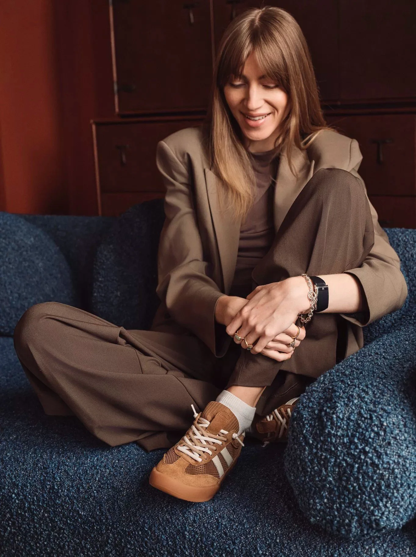 A woman sitting with her legs crossed on a dark blue textured sofa, smiling with eyes closed, in a warm-toned room with wooden furniture.