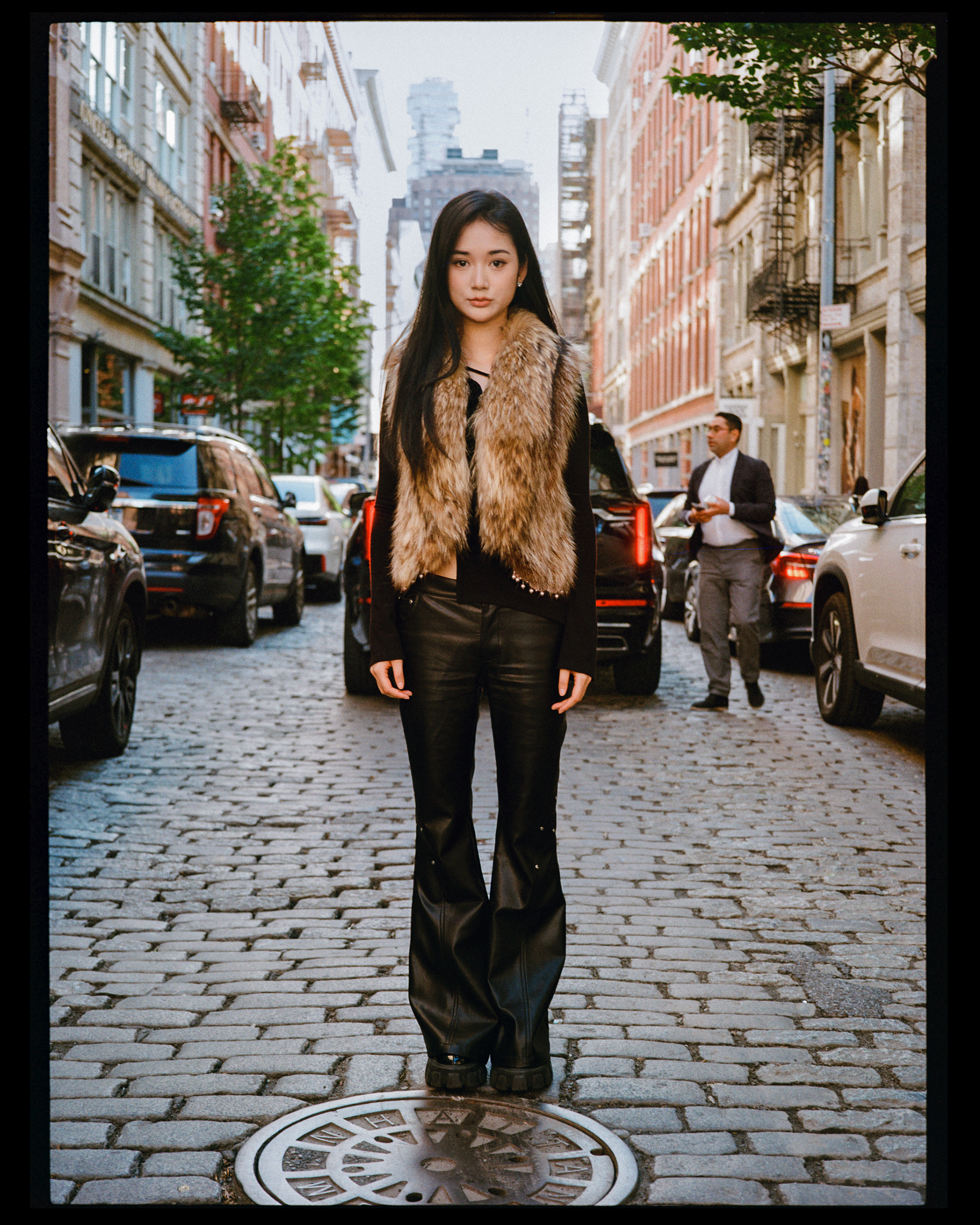 A young woman with long black hair standing on a cobblestone city street, wearing black leather pants, a fur vest, and a black top, with modern buildings lining the street and a man in a suit walking behind her.
