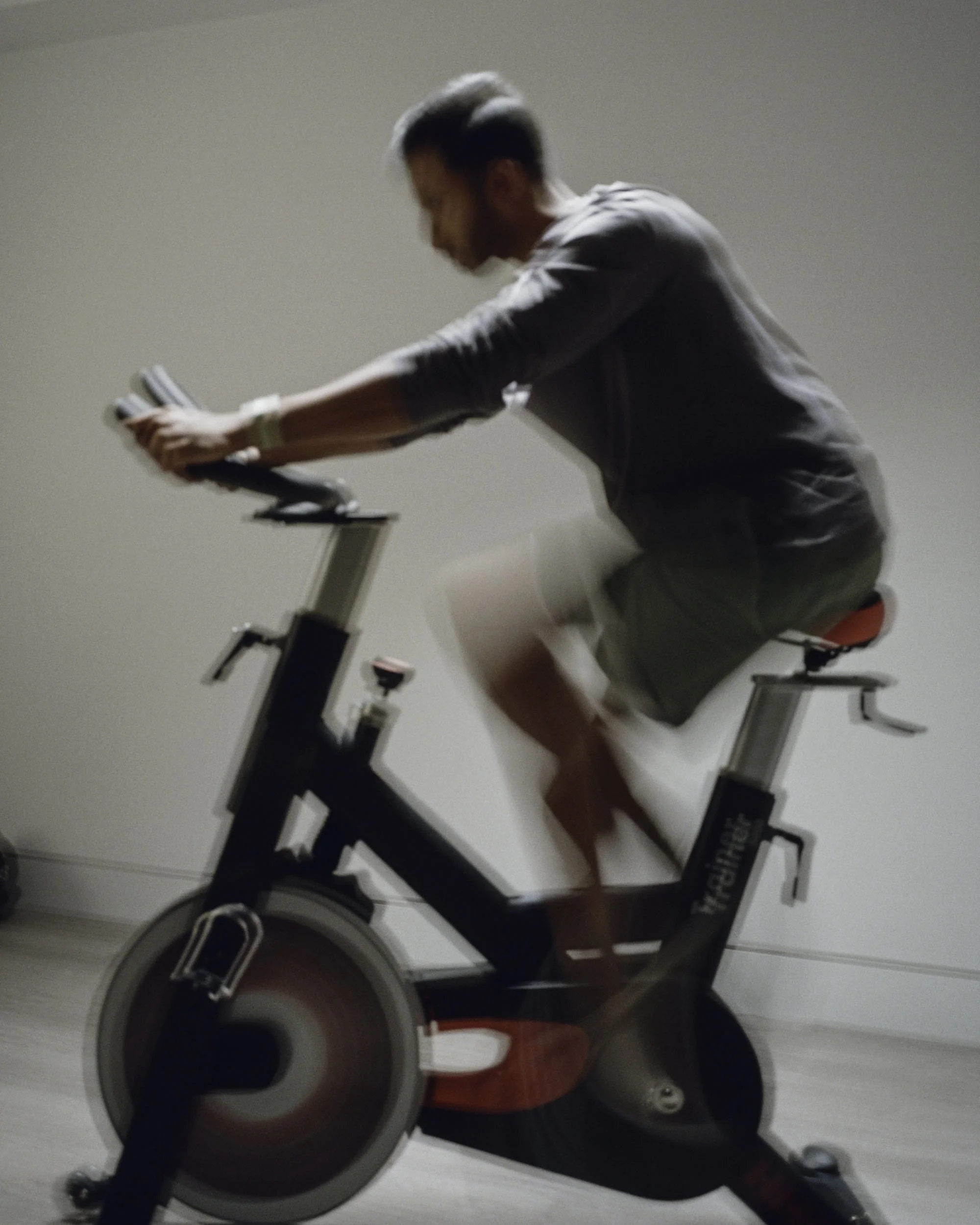 A person with short dark hair, wearing a gray t-shirt, green shorts, black sneakers, and red socks, is sitting on a spinning bike in a room with beige walls. The person is leaning forward with their hands gripping the bike's handles, and they are looking downward.