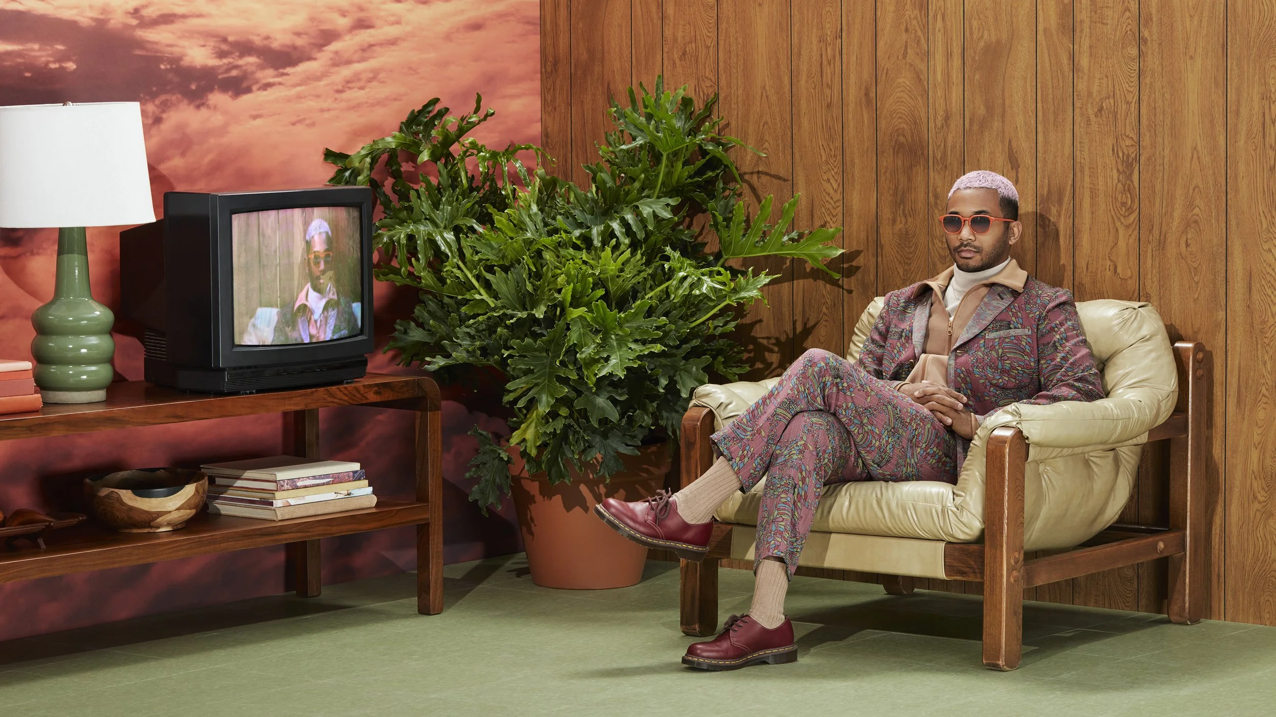 A man with pink hair, sunglasses, and patterned suit sitting on a beige armchair in a room with wooden walls and a large green plant, with a vintage TV on a wooden table displaying his reflection.
