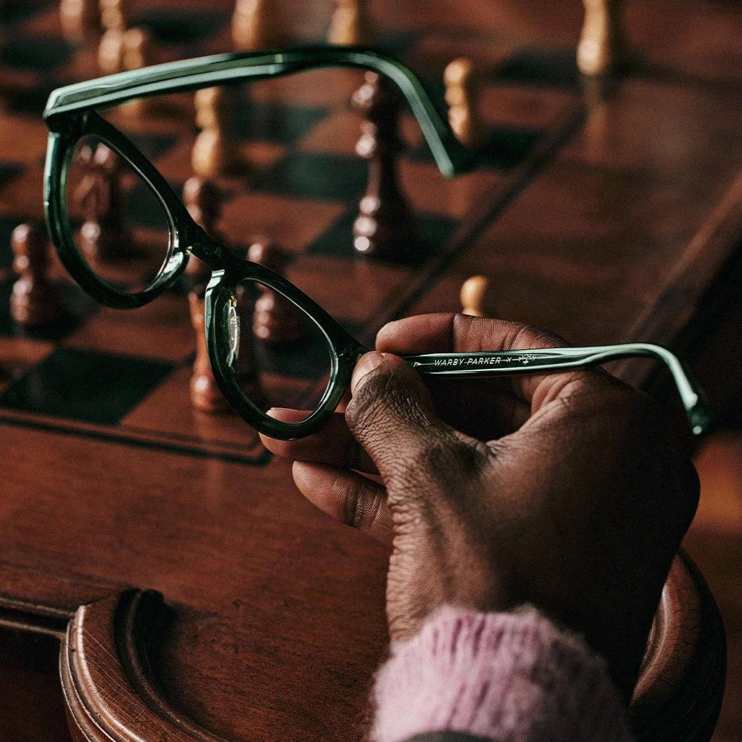 A person's hand holding a pair of black and green Warby Parker eyeglasses over a wooden chessboard with chess pieces in the background.