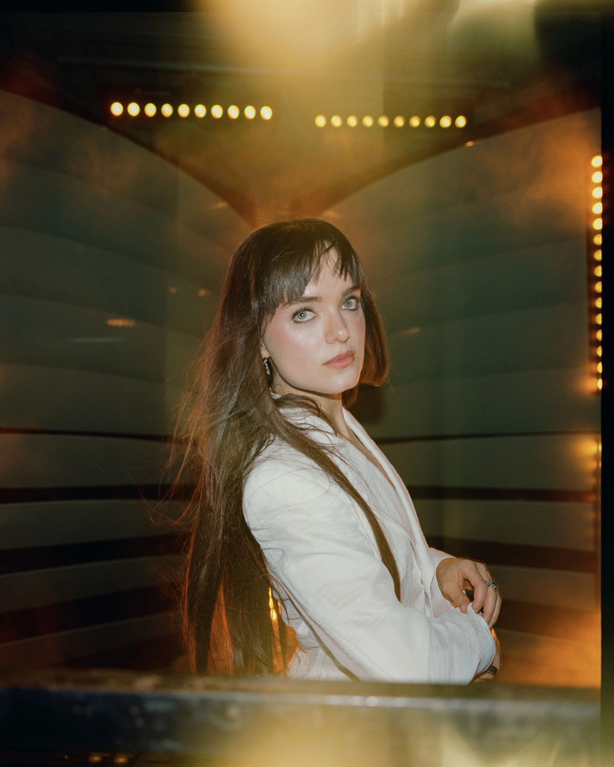A woman with long brown hair and blue eyes is looking at the camera with a neutral expression. She is wearing a white top and earrings, and her arms are crossed in front of her. The background features a metallic or reflective surface with warm yello